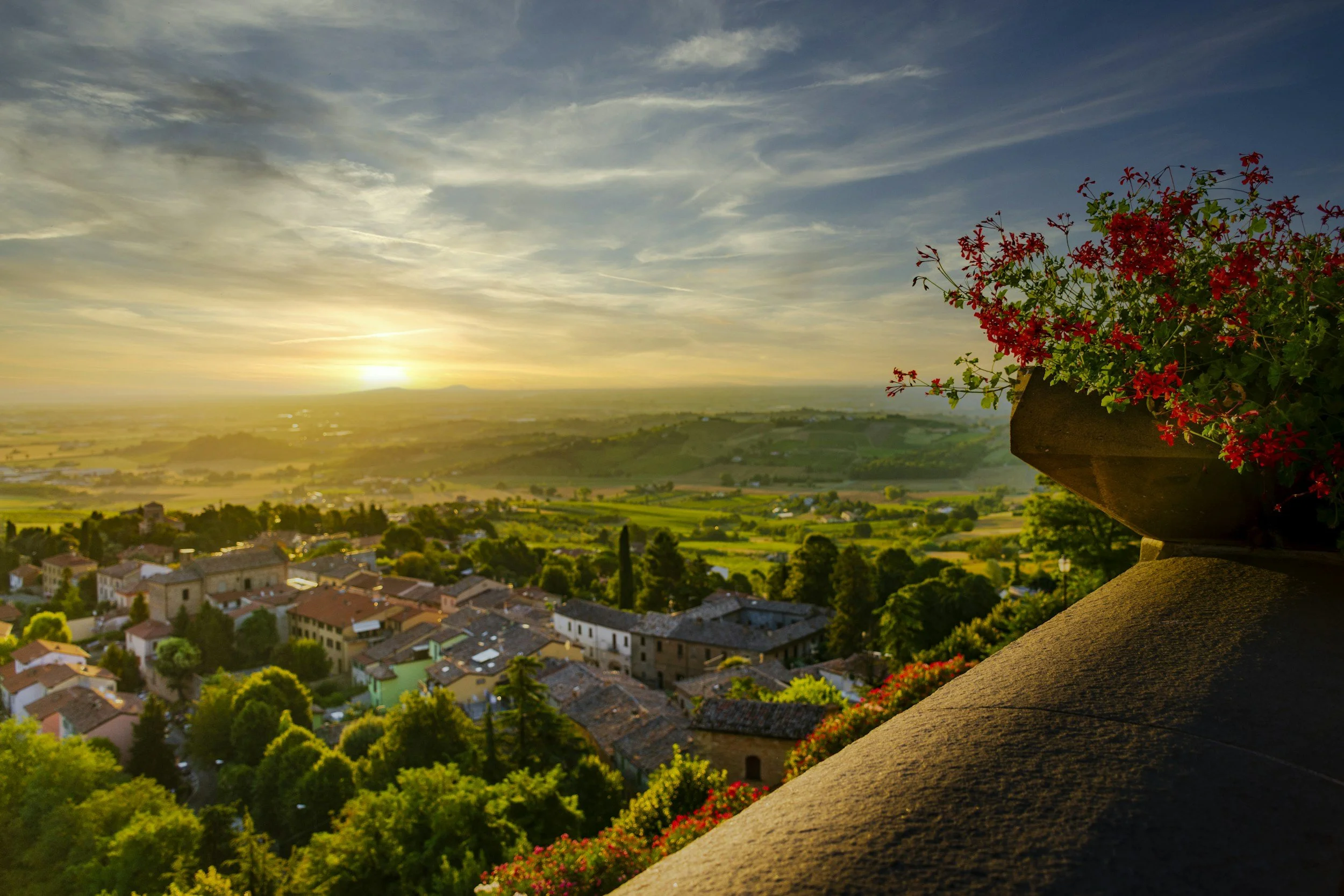 View of a scenic landscape during sunset, with a small town and green fields in the distance, and a balcony with red flowers in the foreground.