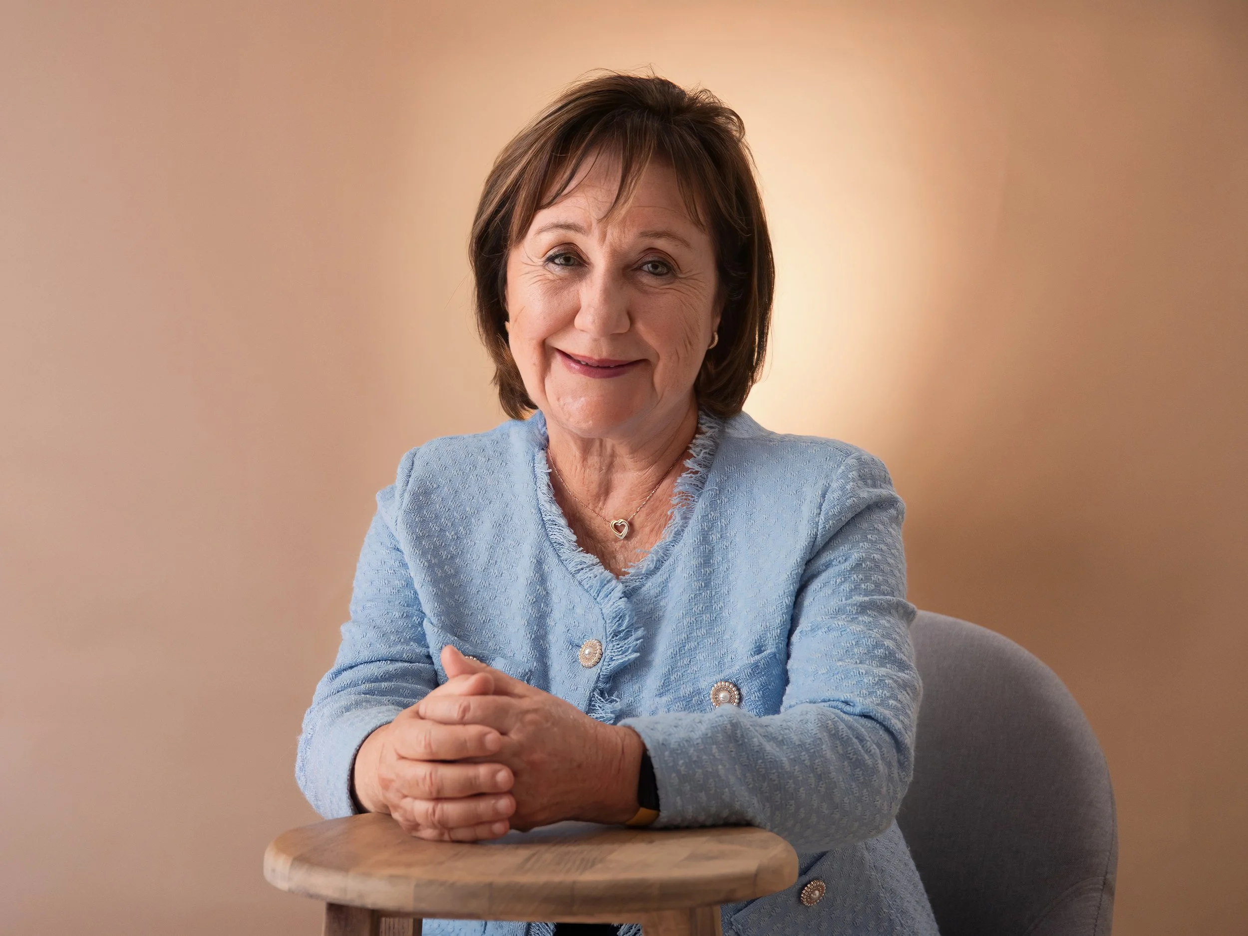 A smiling older woman with short brown hair, wearing a light blue textured jacket, sits at a wooden table with her hands clasped, against a warm beige background.
