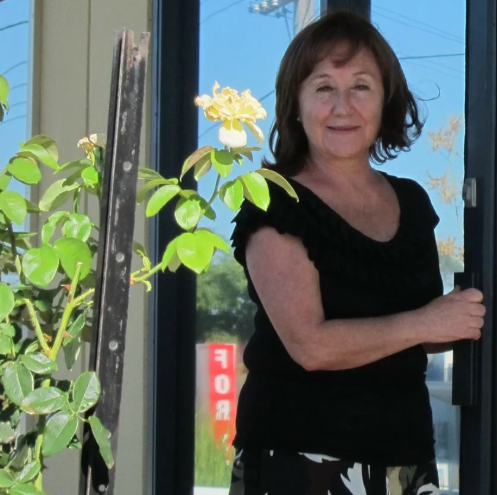 A woman standing outside near a glass door, holding the door handle, with a potted plant with yellow flowers nearby.