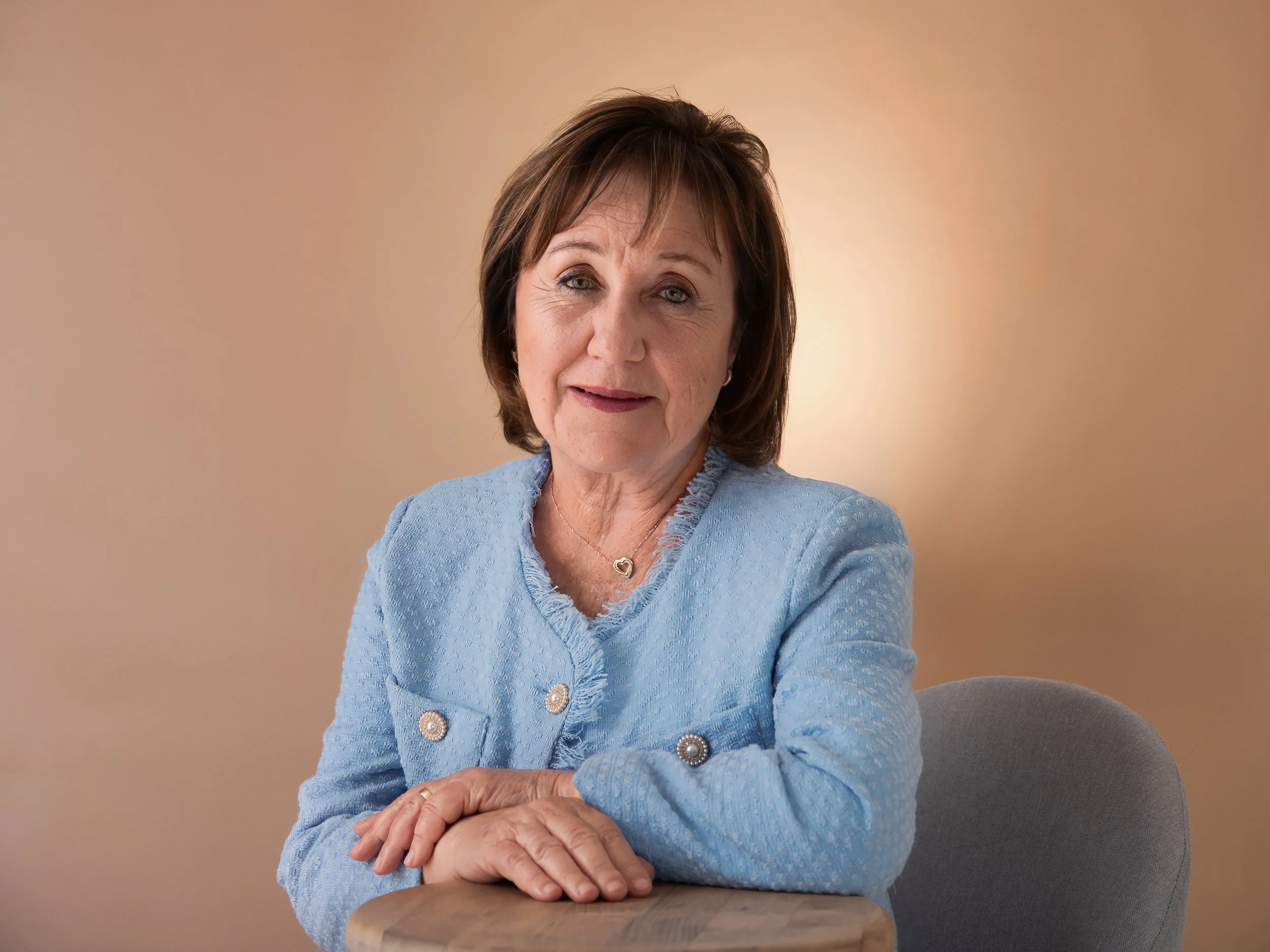 A mature woman with short brown hair, wearing a light blue blazer with decorative buttons, sitting at a table with her arms crossed, looking at the camera with a slight smile against a beige background.