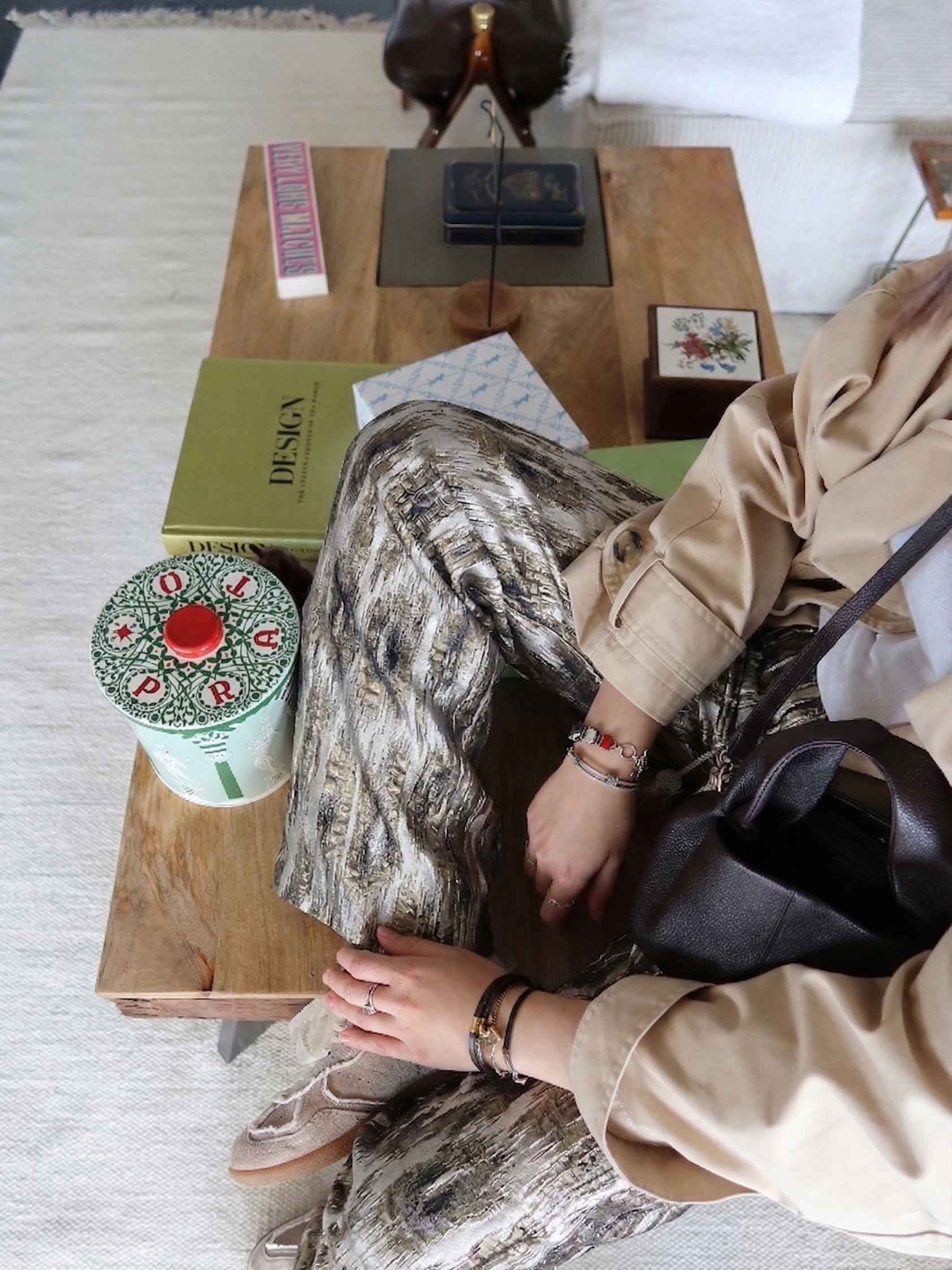 A person sitting at a wooden table with decorative items, a book titled 'DESIGN', a container decorated like a Christmas tree, and various accessories on their wrist, wearing patterned pants and beige jacket.