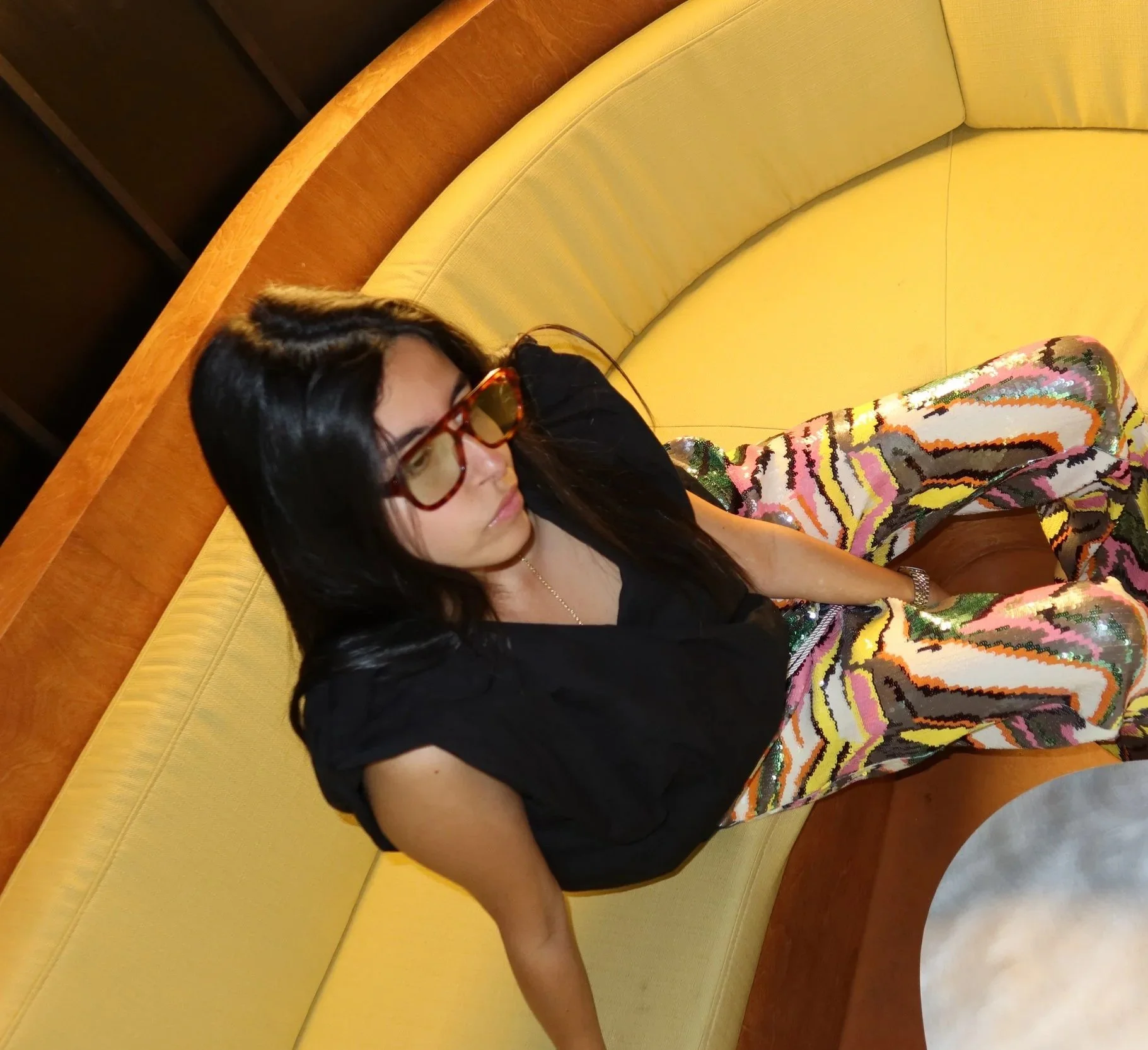 A woman sitting on a yellow cushioned booth in a restaurant, wearing sunglasses and a colorful abstract-patterned skirt, with three glasses of orange-colored drinks on the table in front of her.