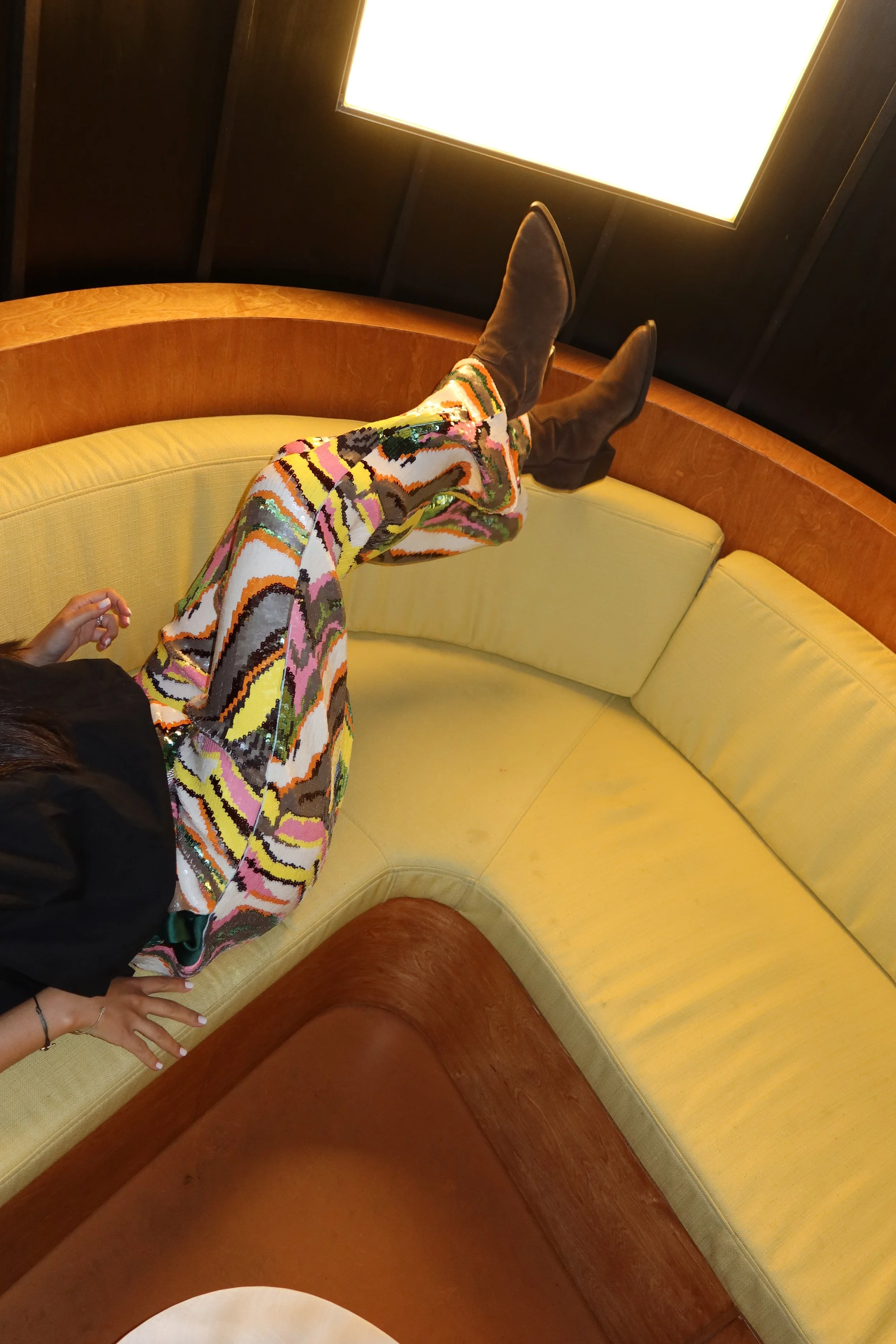 Person lying on yellow cushioned booth seating, wearing colorful, patterned pants and brown boots. The scene appears to be indoors with dark wooden walls and a bright rectangular light fixture above.