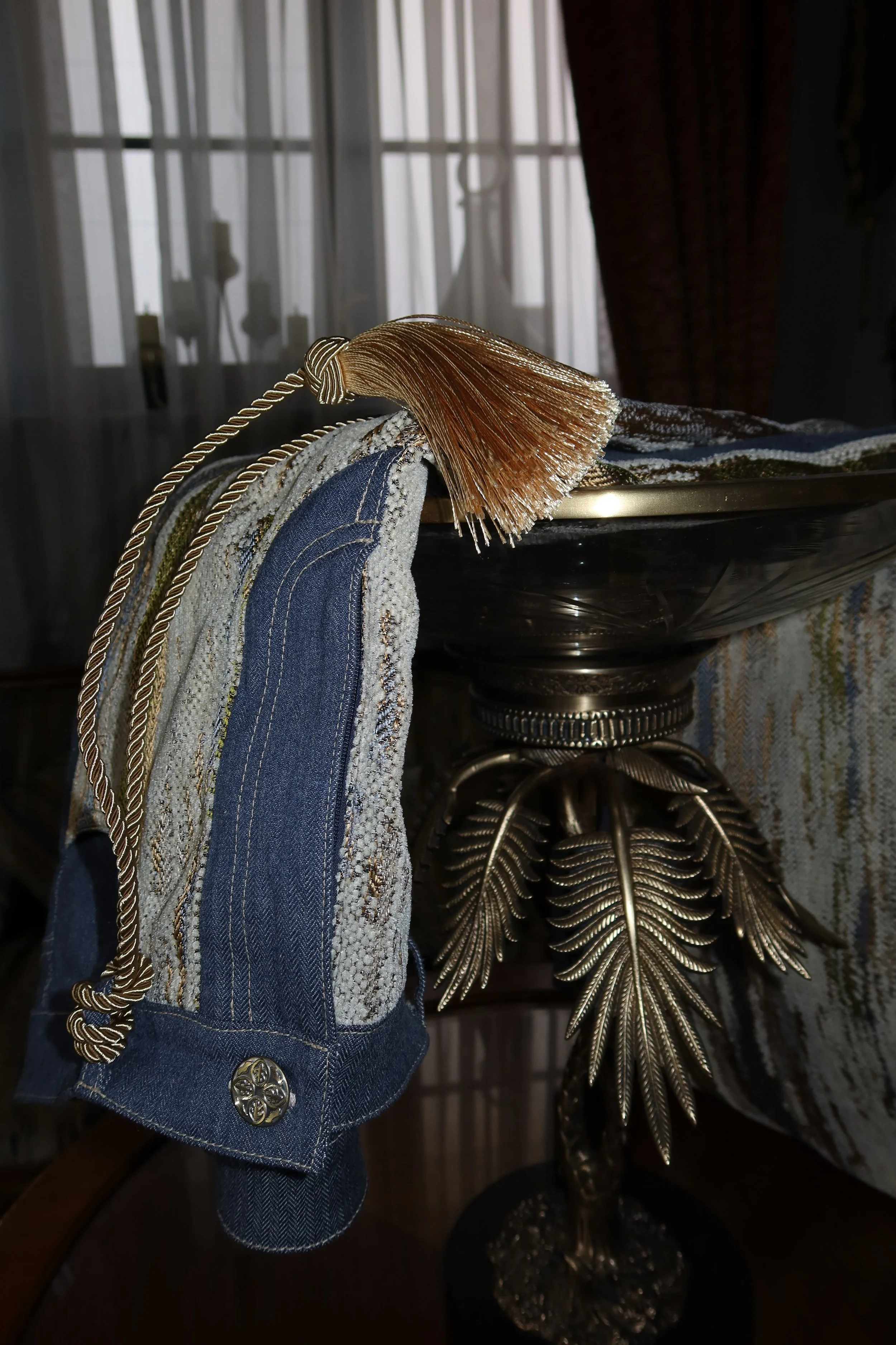 Close-up of a decorative cloth napkin or towel with denim pattern and button, draped over a metallic dish on a dark wooden surface, with a metallic leaf-shaped ornament and a window with sheer curtains in the background.