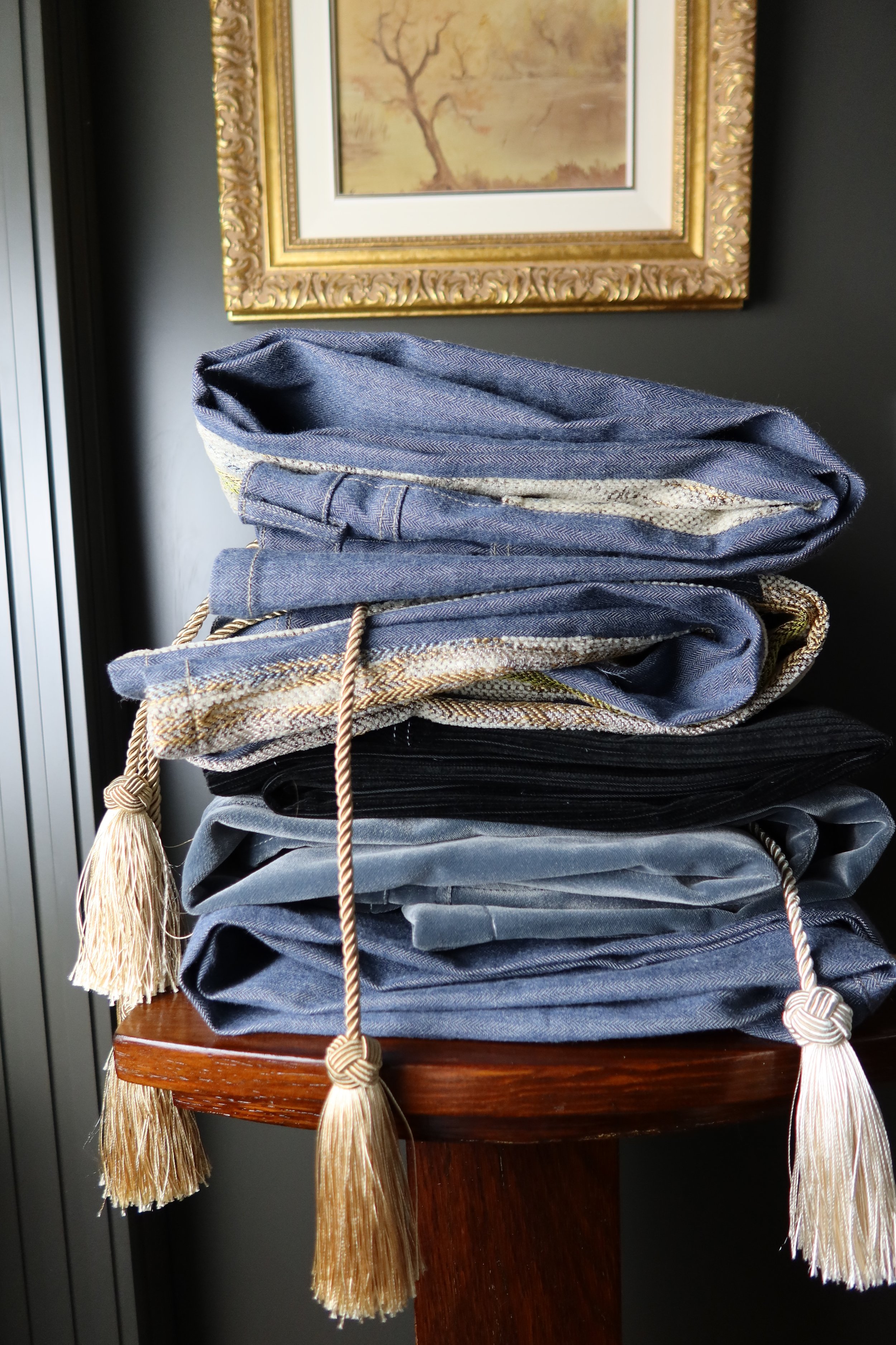 A stack of folded blue and gray clothing, secured with decorative tassels and cords, placed on a wooden stand in front of a framed painting of a tree. fun pants, fun jeans, summer jeans, funky pants