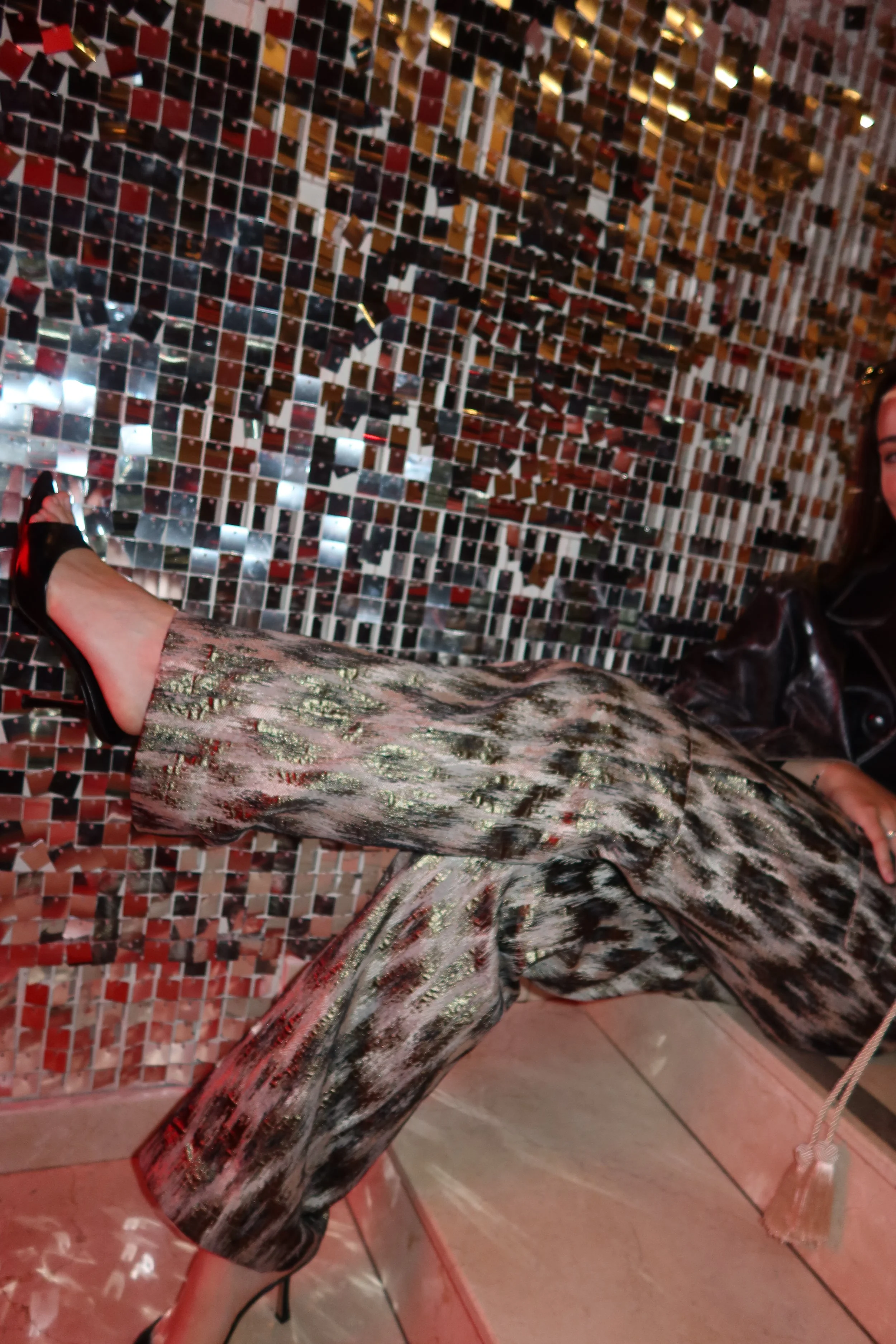 Person wearing black sandals, patterned pants, and a shiny, metallic dress with a pink and silver pattern, standing in front of a wall decorated with small reflective tiles.