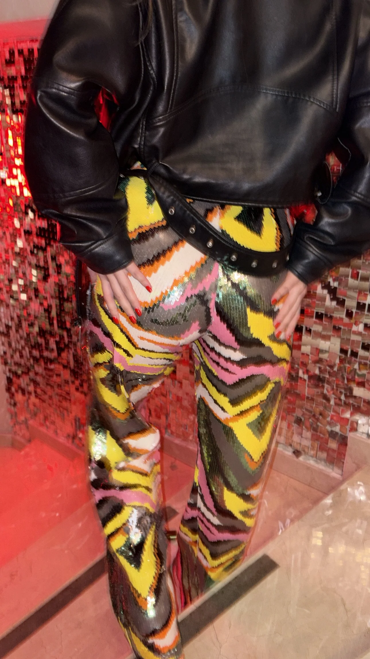 A person wearing a black leather jacket, colorful zebra print pants with yellow, pink, and black patterns, and red nail polish. They are standing in front of a red mosaic wall.