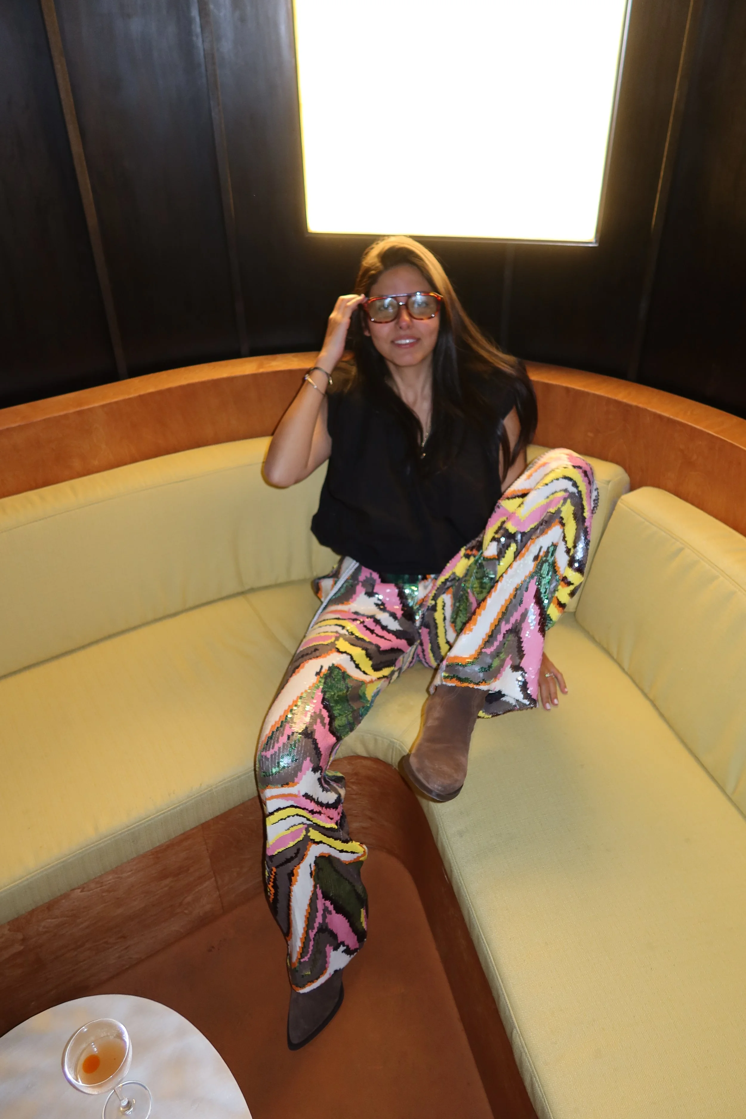 Woman with long dark hair wearing glasses, a black top, patterned pants, and boots, sitting on a yellow curved sofa in a dimly lit room with dark walls and a bright square light above her.