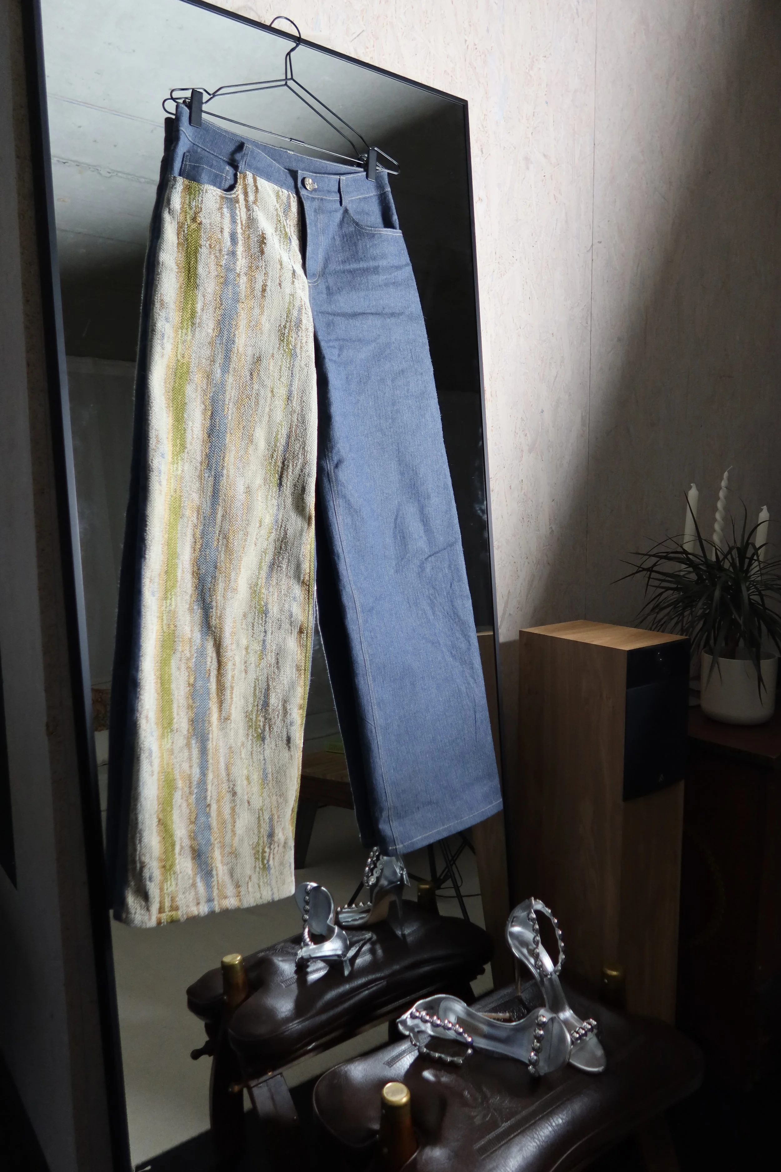 Pair of pants hanging on a hanger in front of a mirror, with one pant leg patterned and the other solid blue, near shelves with shoes and decorative items.