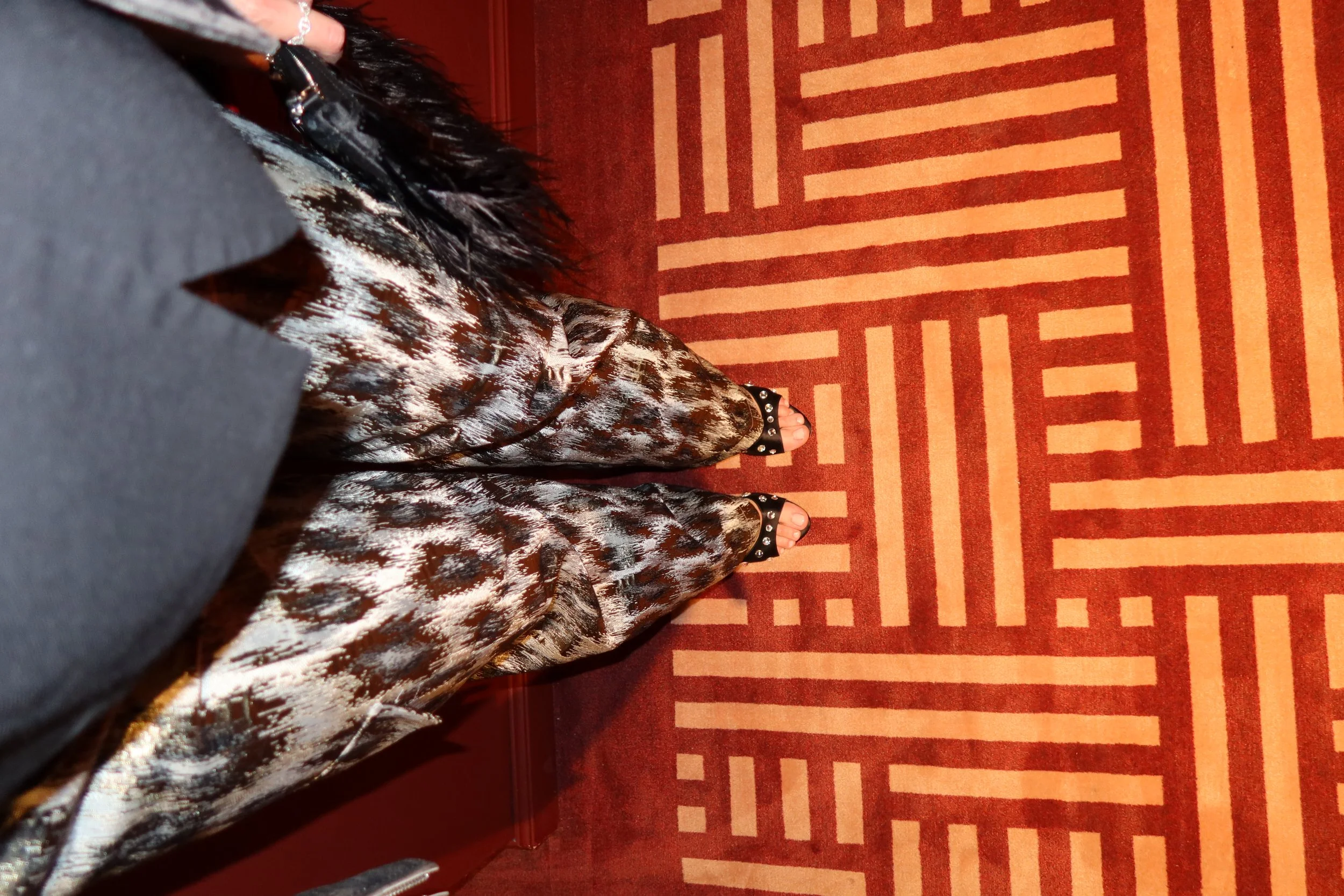 Person standing on a patterned red and beige carpet, wearing open-toed sandals with embellishments and patterned pants.