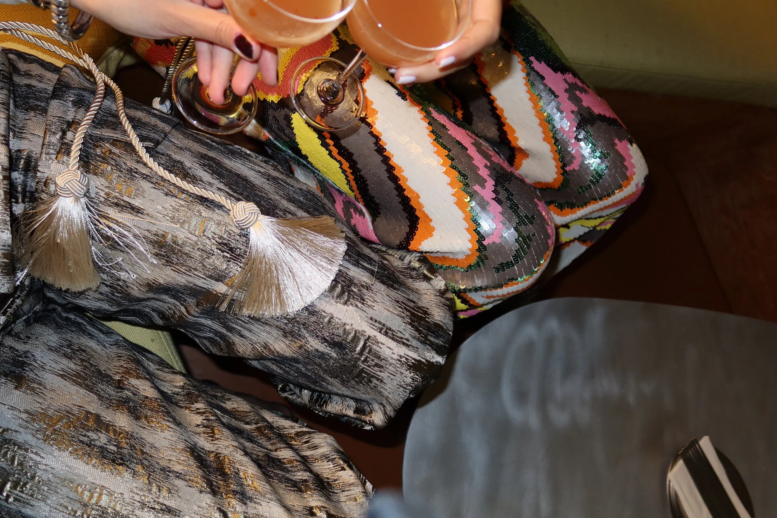 Two hands holding wine glasses filled with a light pink cocktail, resting on a colorful, shimmering, sequined fabric with black, pink, orange, and white stripes and intricate patterns.