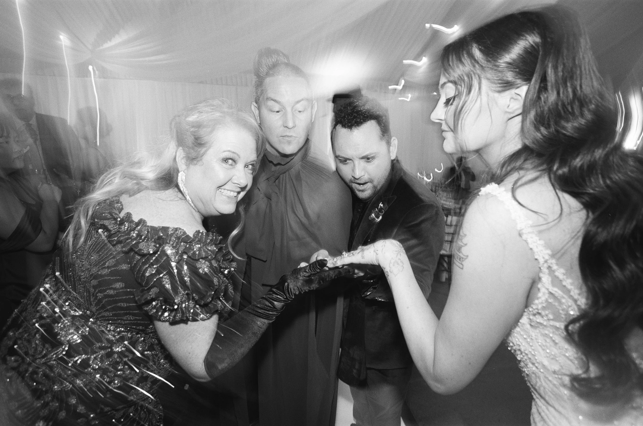 Four people at a party, one woman smiling and showing her teeth, wearing a sequined dress and gloves, all looking at the camera. The three men and women are closely gathered, with one man holding something in his hand, creating a lively, energetic sc