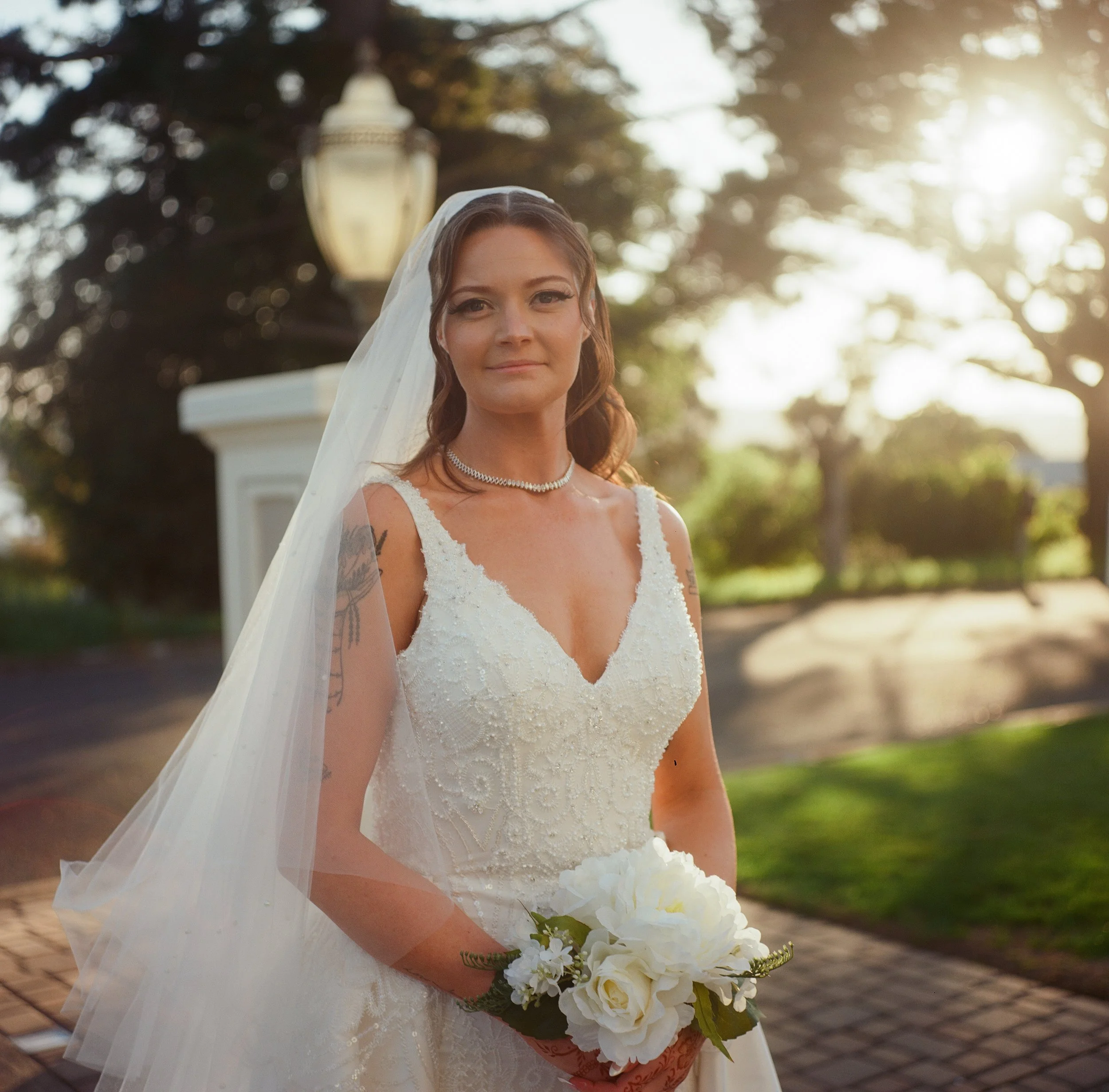 A bride in a white wedding dress holding a bouquet of white flowers, standing outdoors at sunset, with trees and a lamppost in the background.