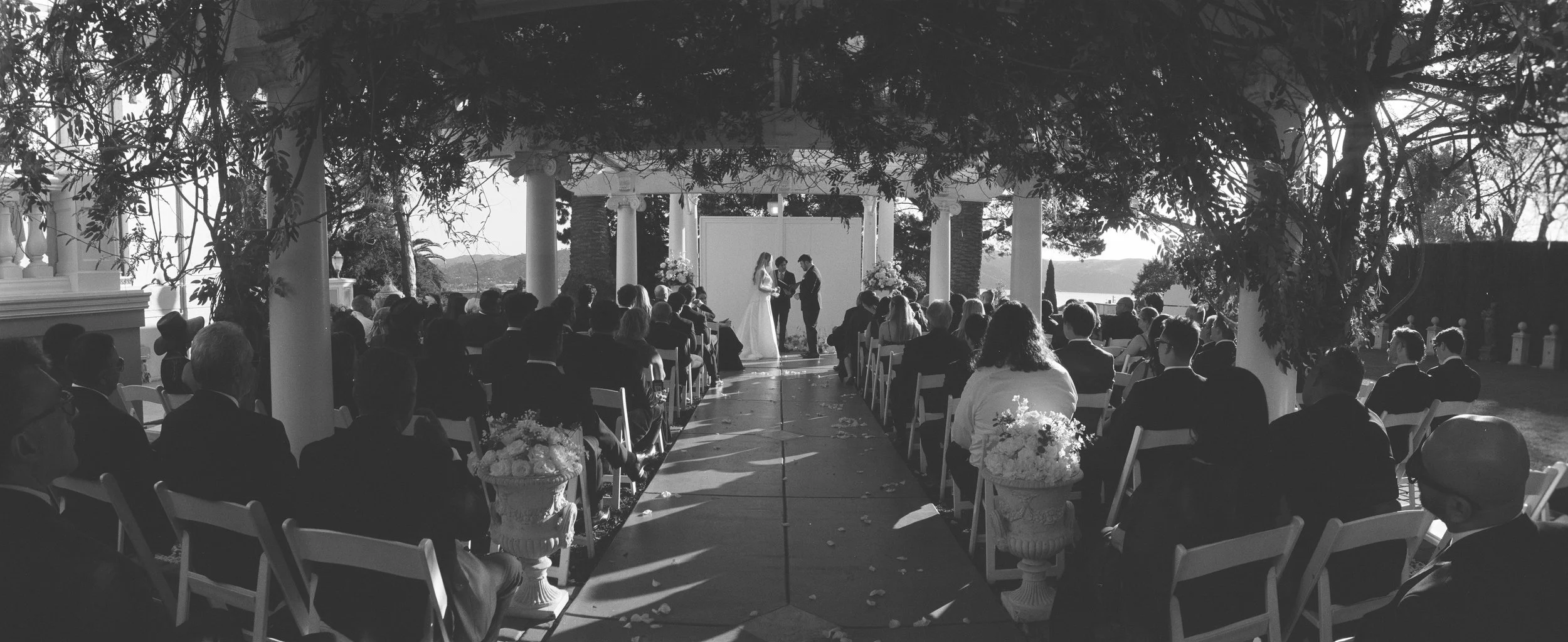 A black and white photo of an outdoor wedding ceremony with guests seated under trees and a view of water in the background. The couple is exchanging vows at the altar with officiant.