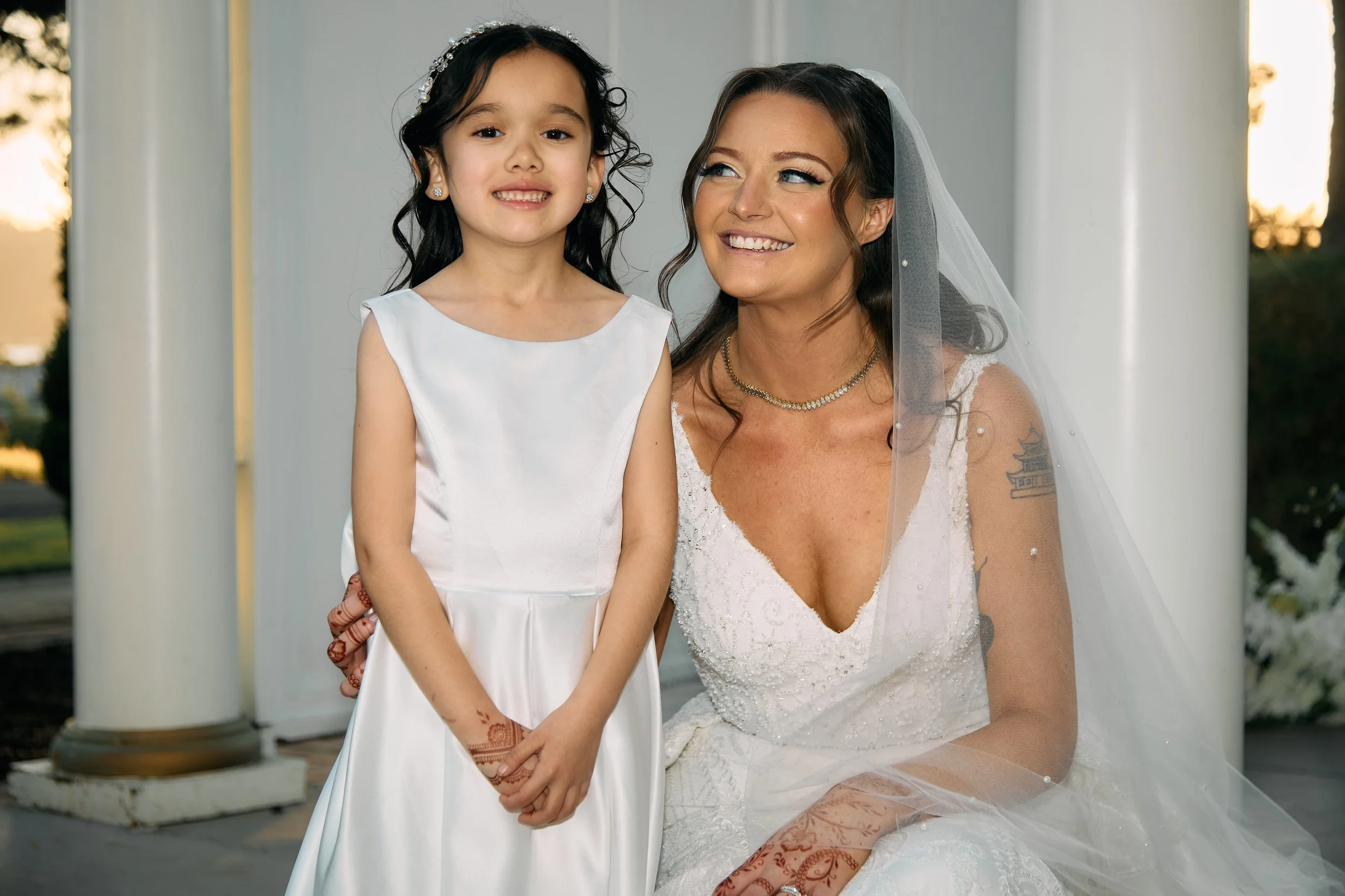A bride and a young girl, possibly a flower girl, smiling outdoors during a wedding, with large white columns in the background.