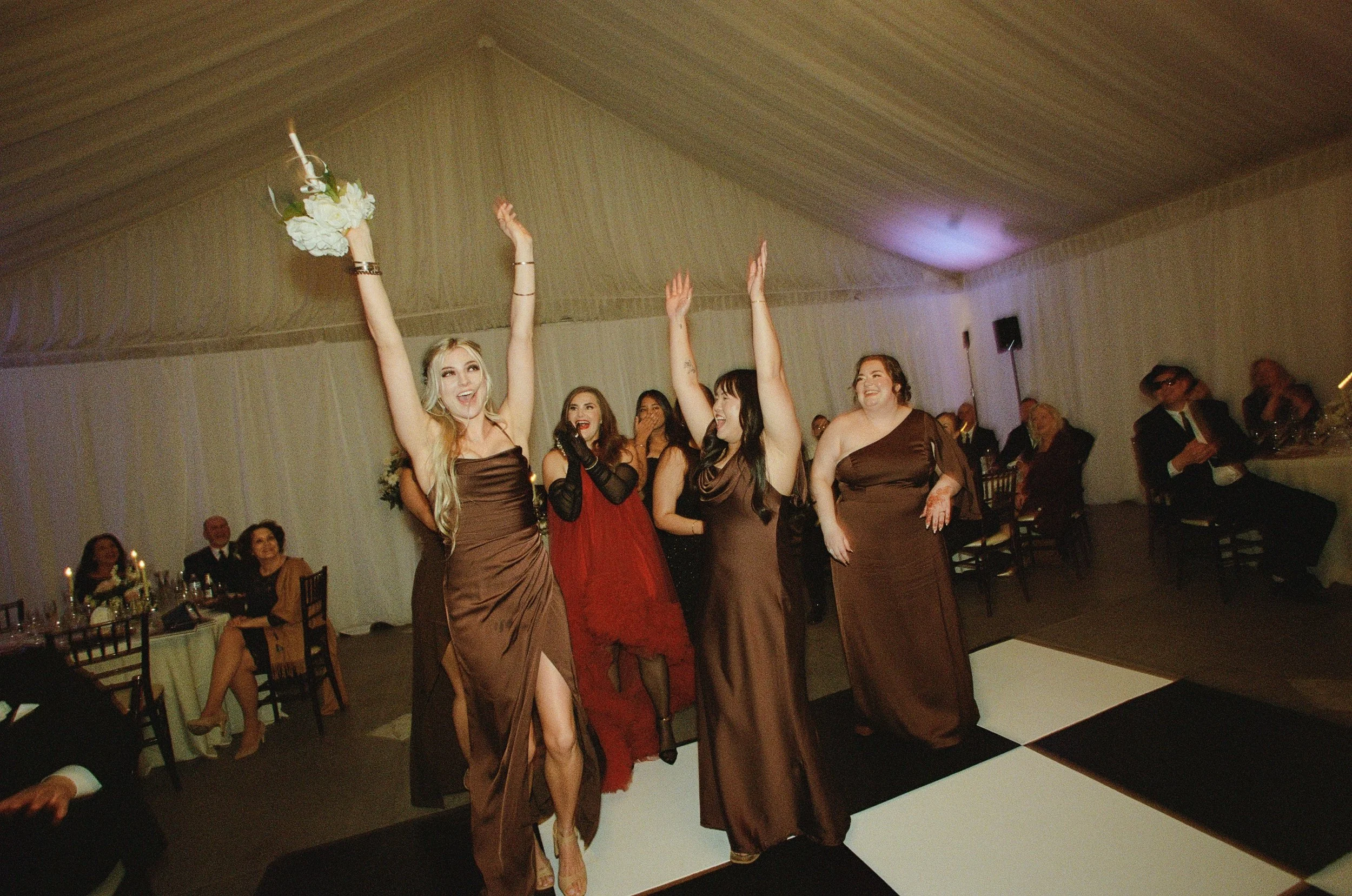 Four women in elegant dresses dancing and celebrating at a wedding reception, with guests seated at tables watching.