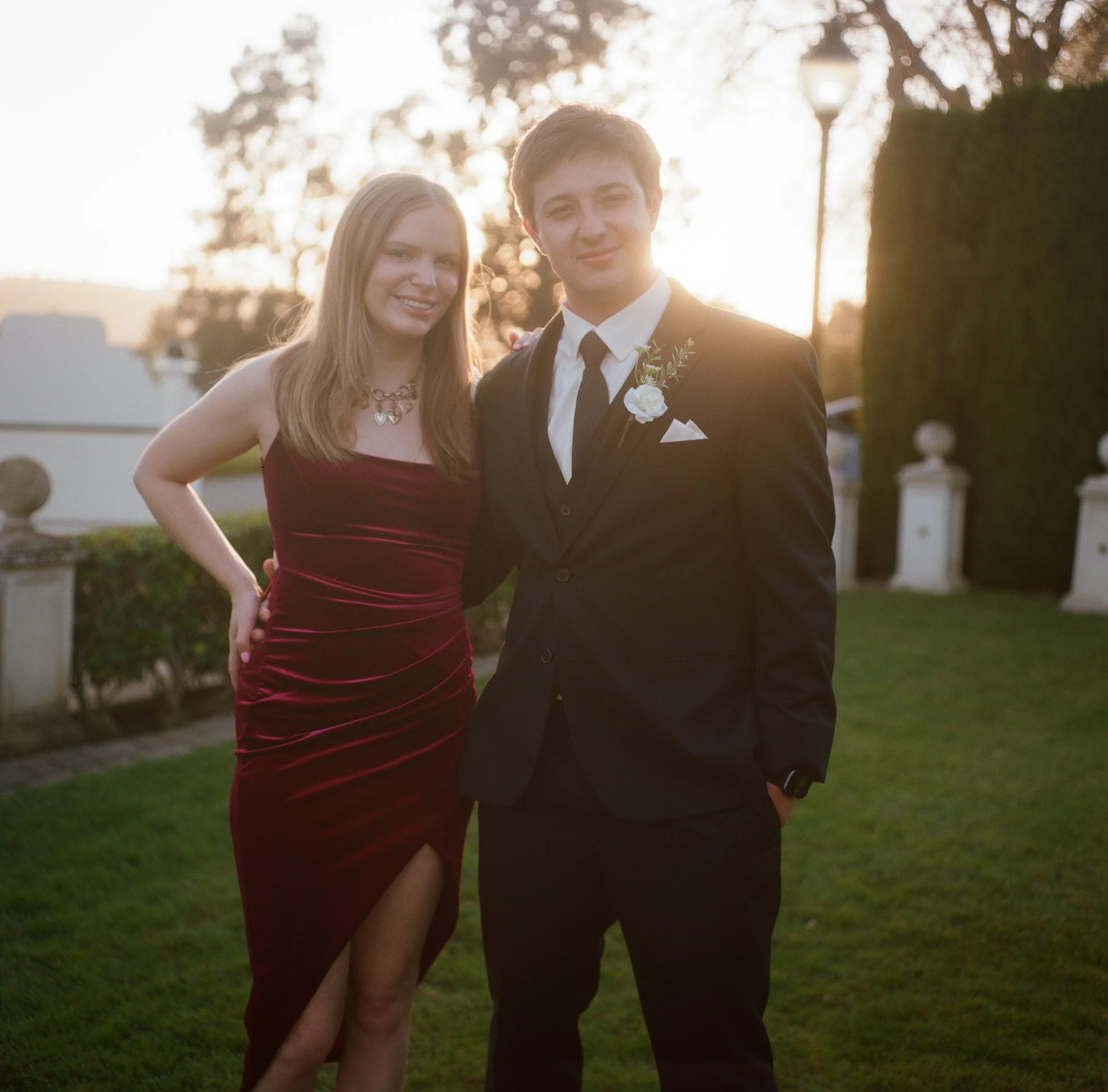 A young woman in a burgundy gown standing next to a young man in a black tuxedo, both smiling outdoors during sunset, with greenery and decorative outdoor elements in the background.