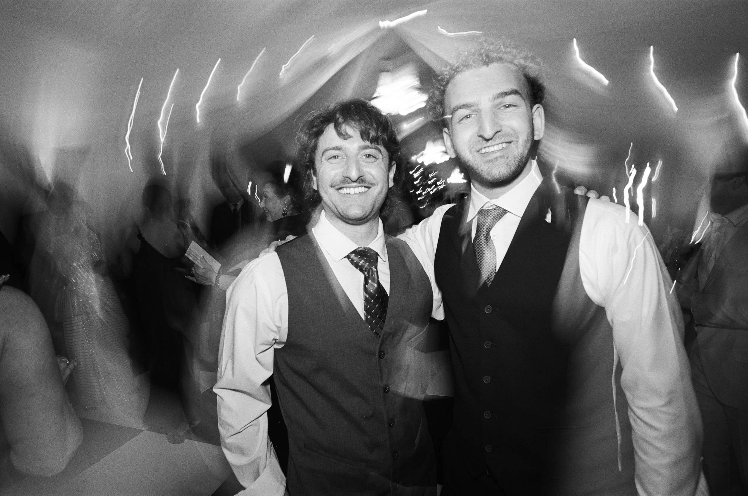 Two men smiling at a party or event, with blurred background of other attendees, some reading, and bright light streaks overhead.