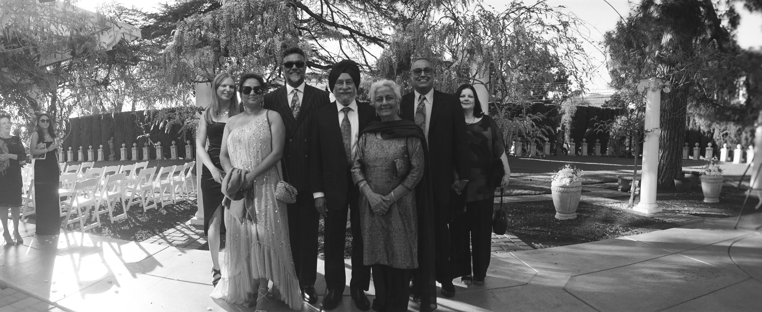 Group of nine people standing outdoors, some dressed formally, posing for a photo in a garden or park with trees and columns in the background