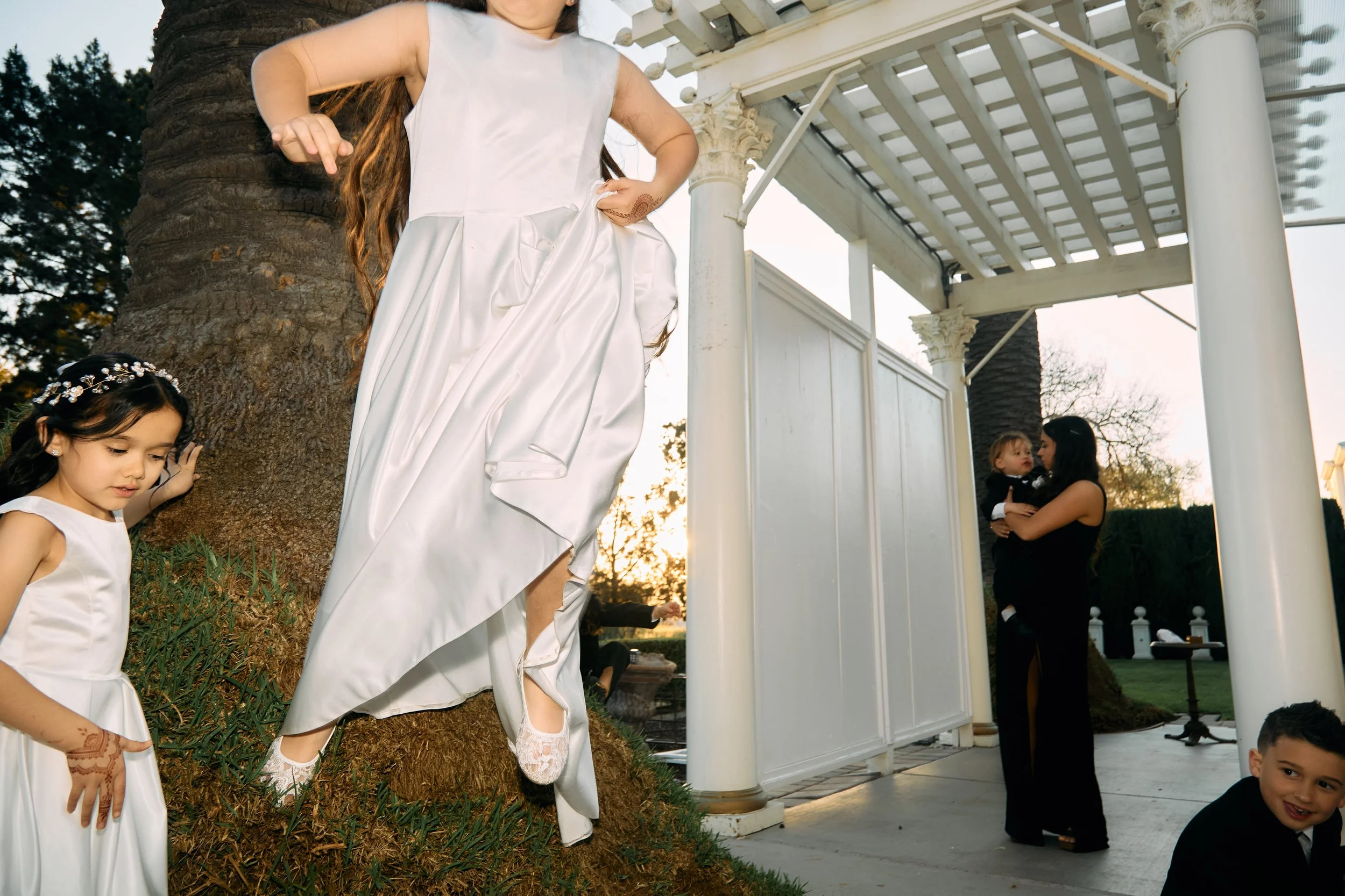 Young girl in a white dress stepping up on a small grassy hill near a large tree during a daytime outdoor event, with another girl in a white dress nearby, a woman holding a child in black attire, and a smiling boy in black sitting on the ground.