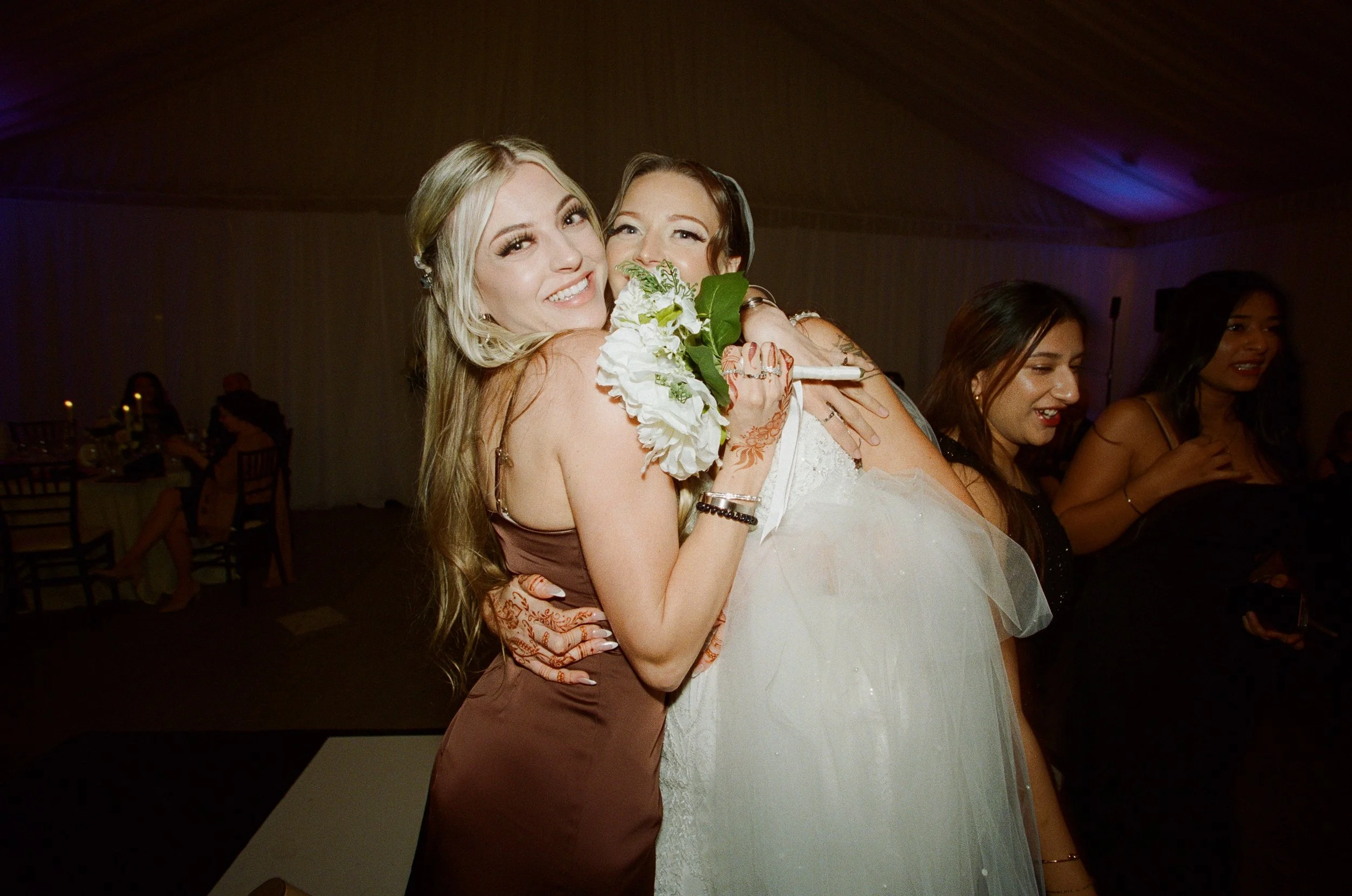Two women, one wearing a brown dress and the other a white wedding gown, hugging at a celebration event, with other women in the background.