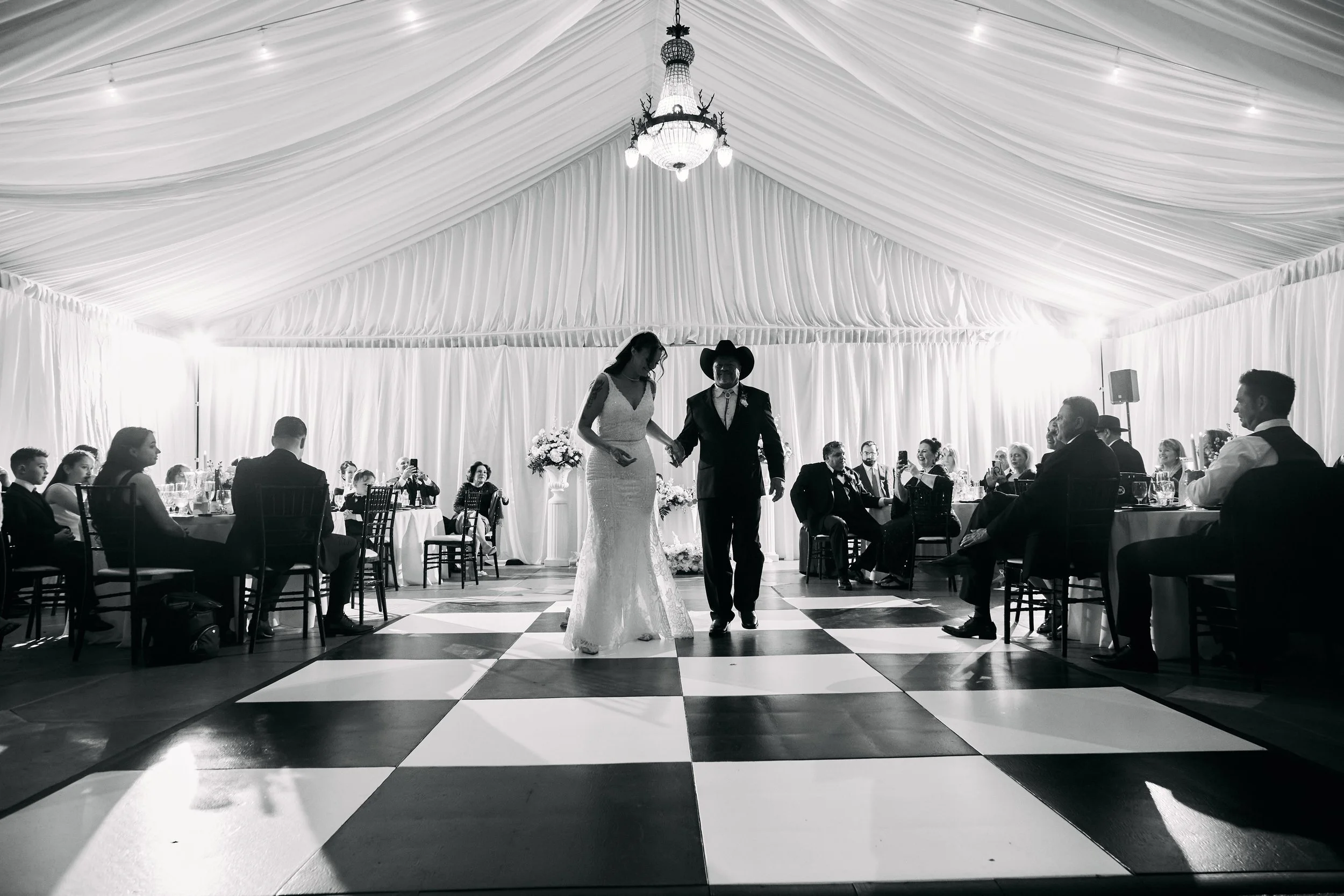 Black and white photo of a bride and groom dancing in a decorated reception tent with guests seated at tables around the dance floor.