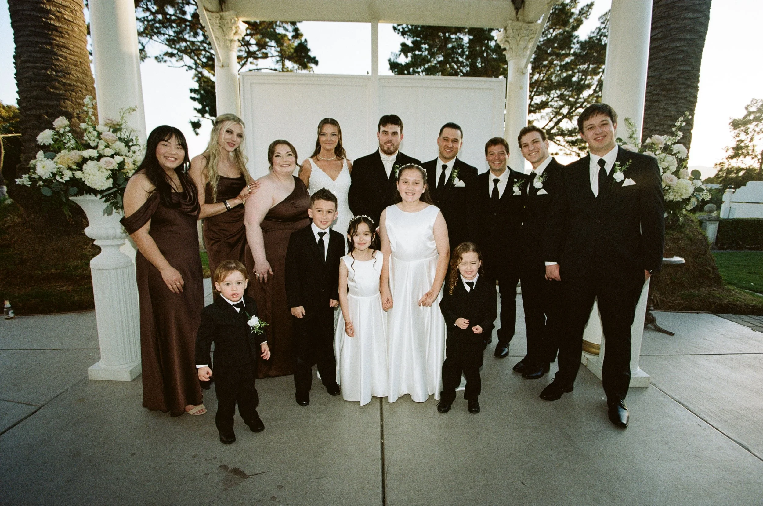 A large group of people celebrating a wedding outdoors, with the bride and groom in the center and family members around them, under a decorative white gazebo with floral arrangements.