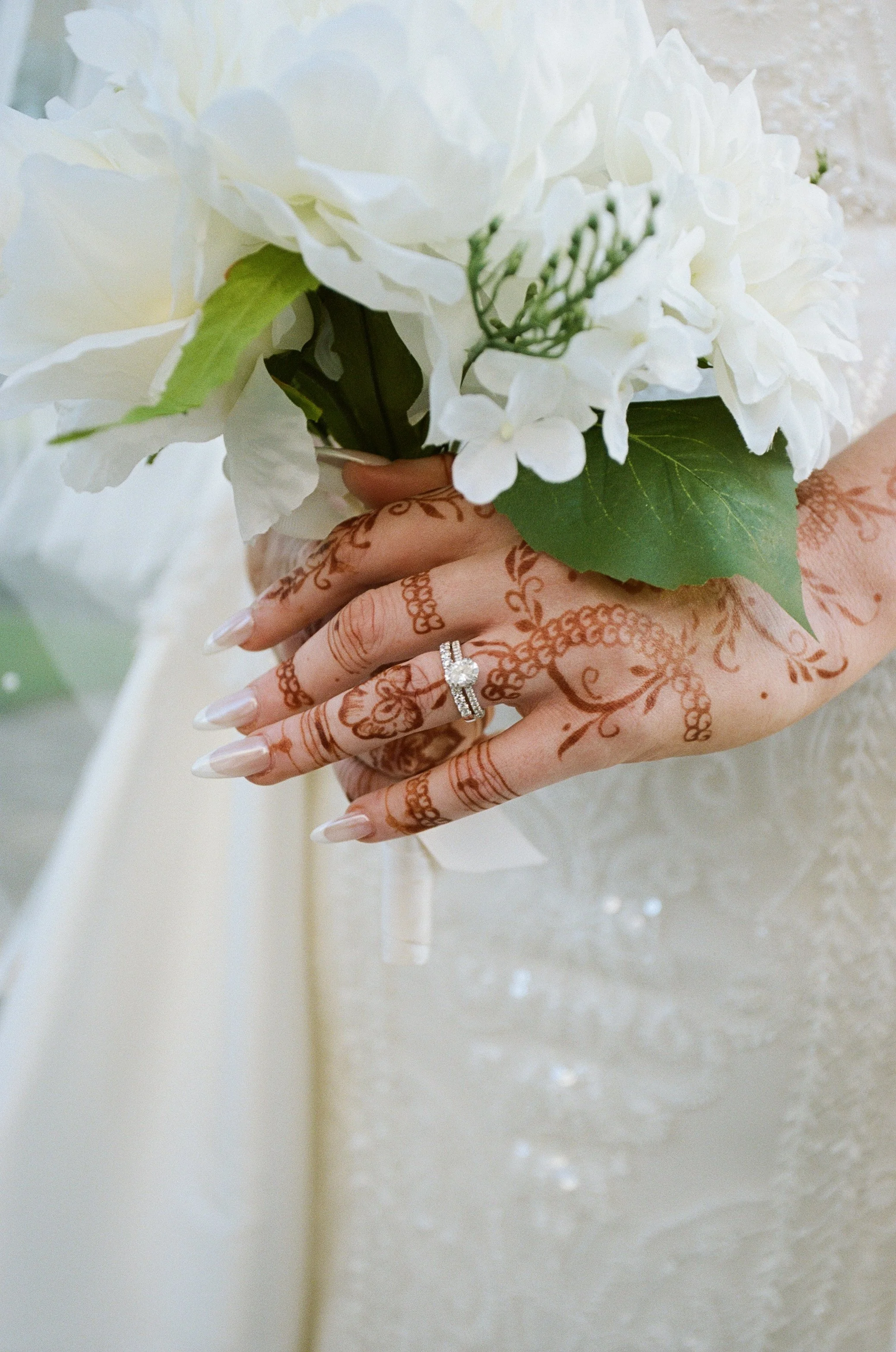 Close-up of a bride's hand holding a white bouquet, showing intricate henna designs on her fingers and a diamond wedding ring.