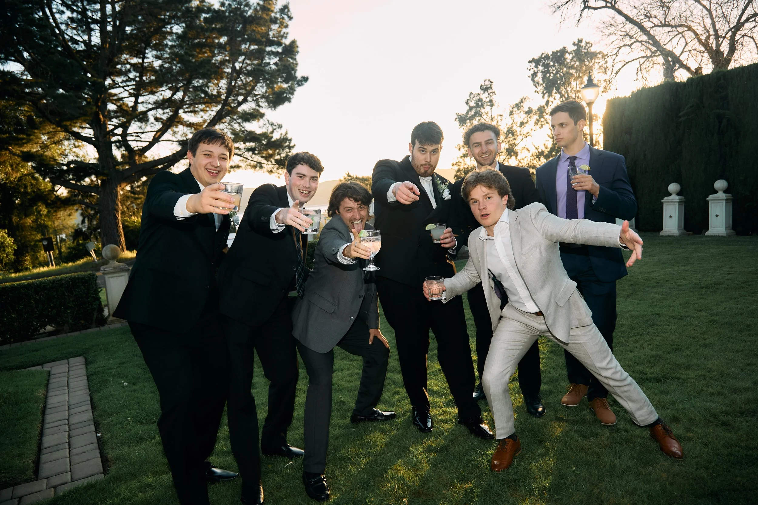 Group of men in suits celebrating outdoors during sunset, holding drinks and smiling.