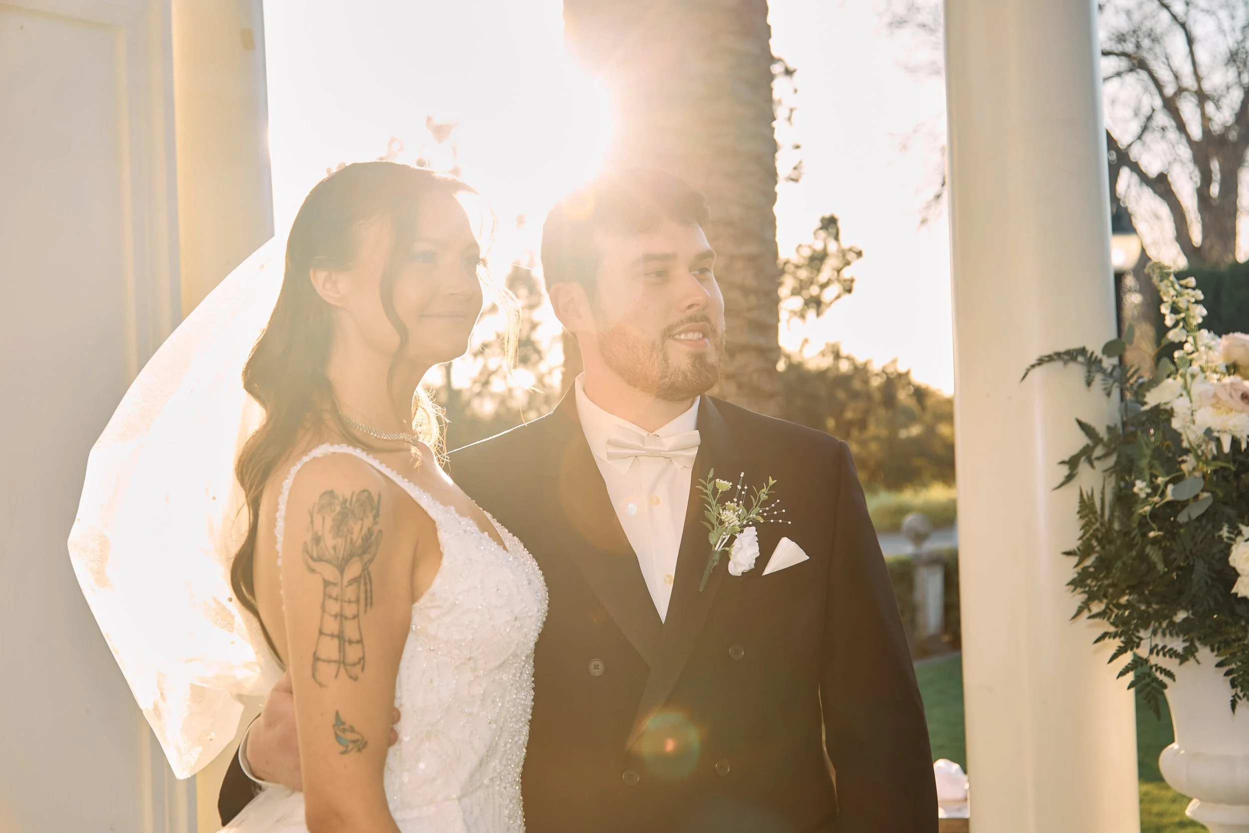 Bride and groom standing outdoors during sunset, wedding ceremony, with columns and greenery in the background.