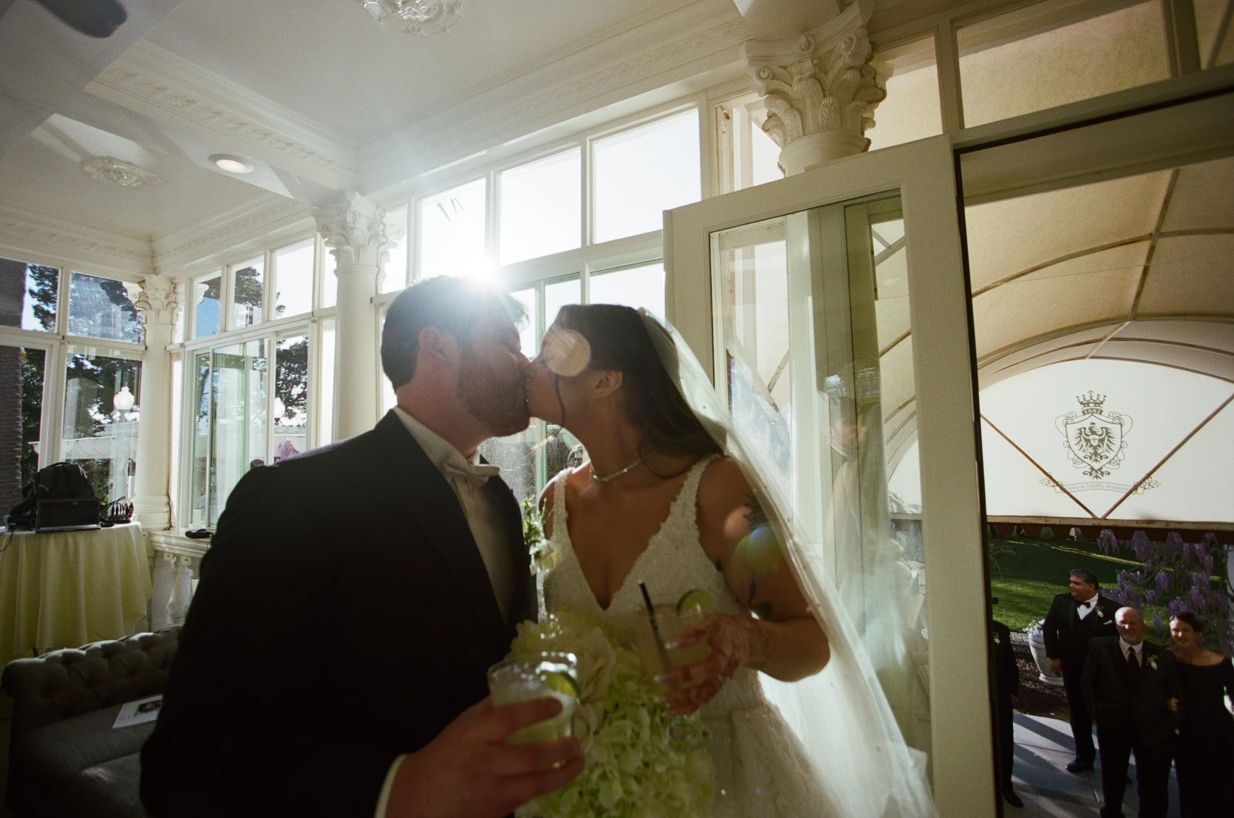 A bride and groom kissing indoors during their wedding, with sunlight shining from behind. The bride is wearing a lace wedding dress and veil, while the groom is in a dark tuxedo. They are holding drinks with lime garnishes.