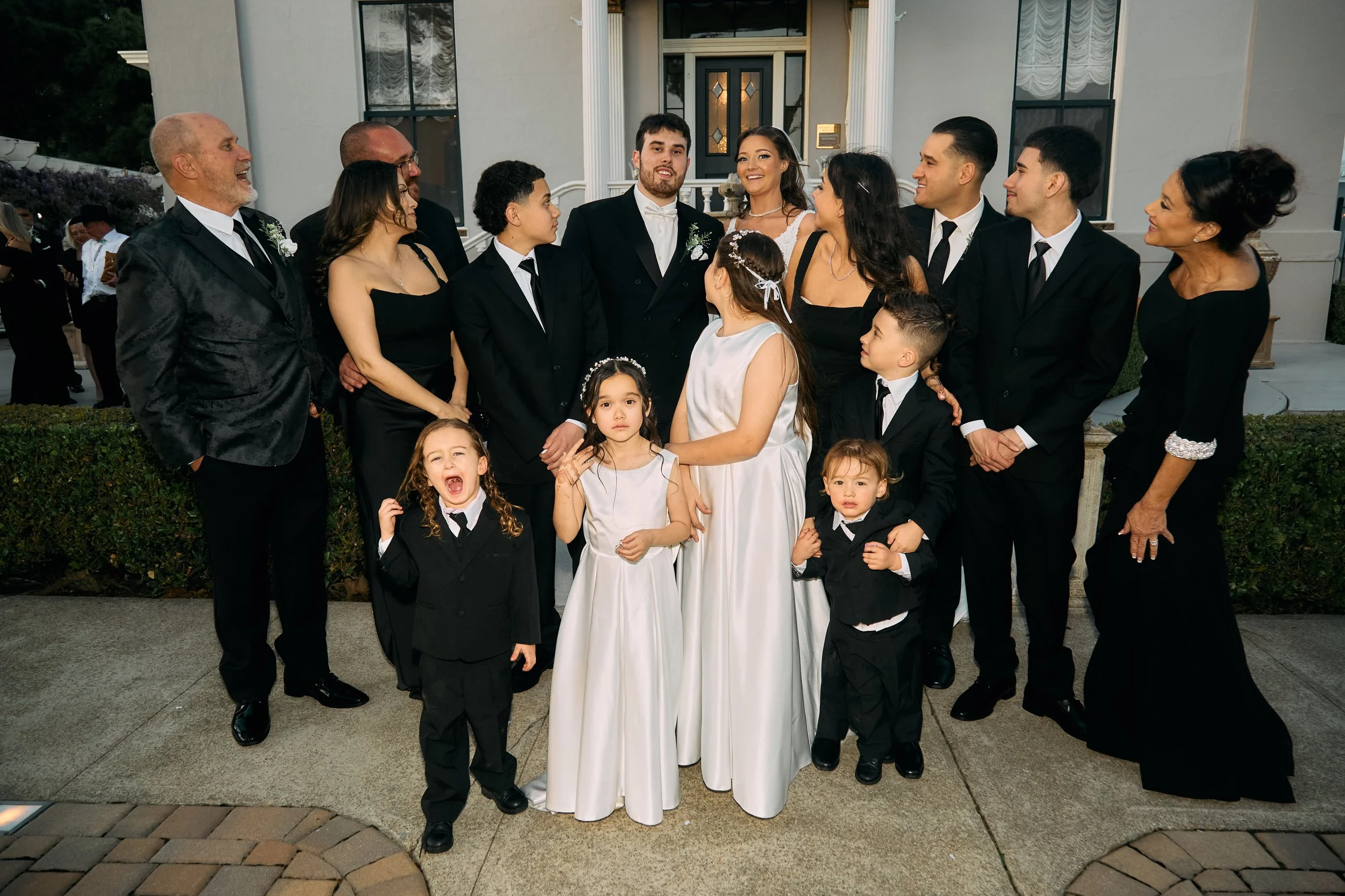 A group of people dressed in formal attire, including a bride and groom, posing outside a house during a wedding celebration.