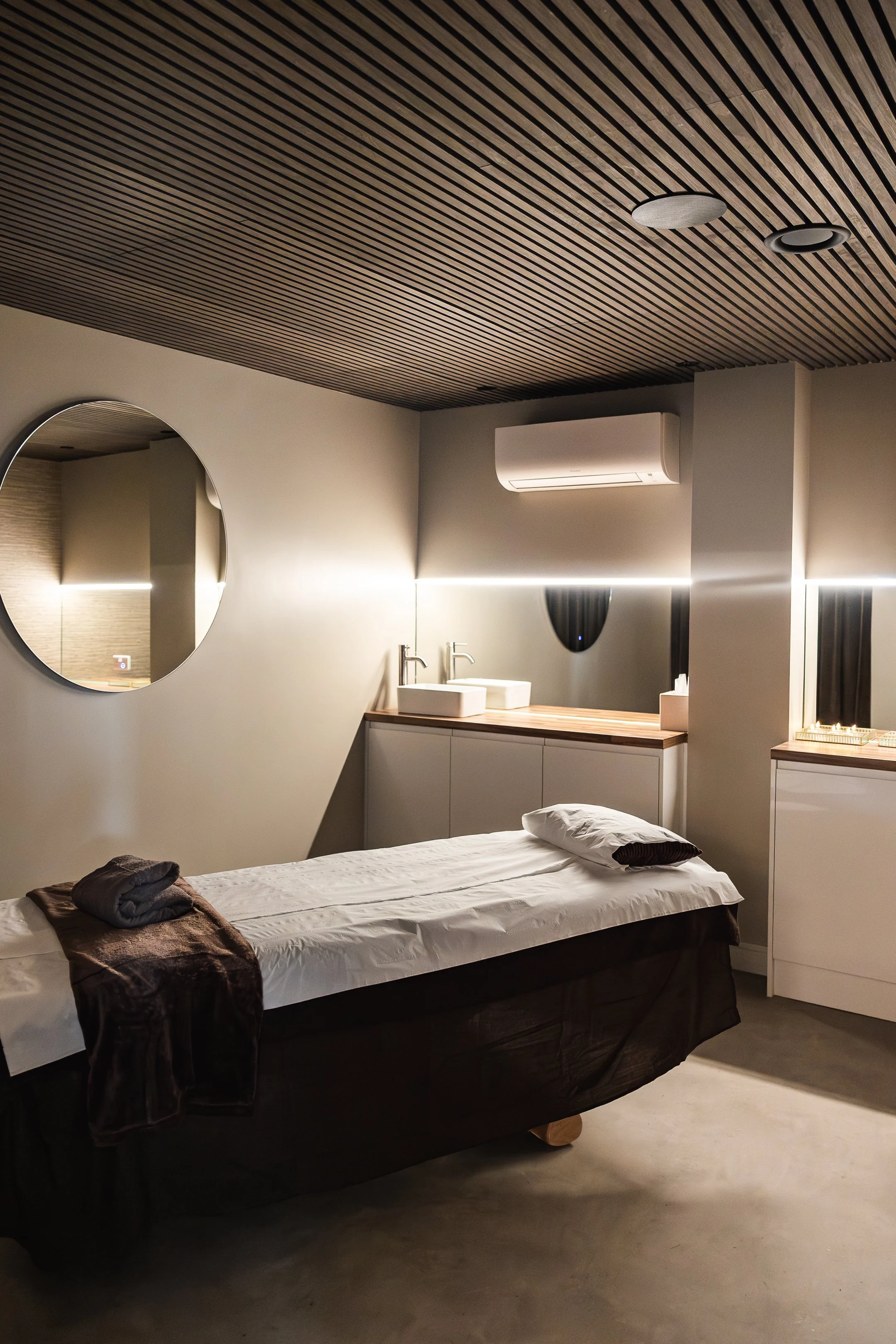 Modern massage room with massage table, pillow, and towel, mirror, sink, and cabinets, with soft lighting and wooden ceiling.