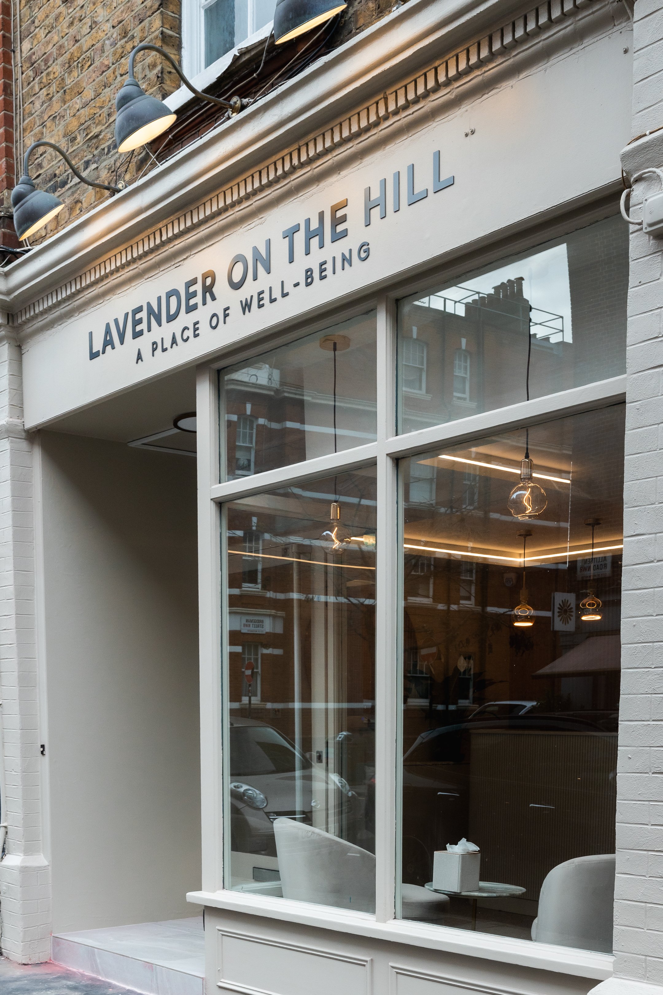 Storefront with a white sign that reads "Lavender on the Hill, A Place of Well-Being" above a large glass window showing the interior with modern pendant lights and seating.
