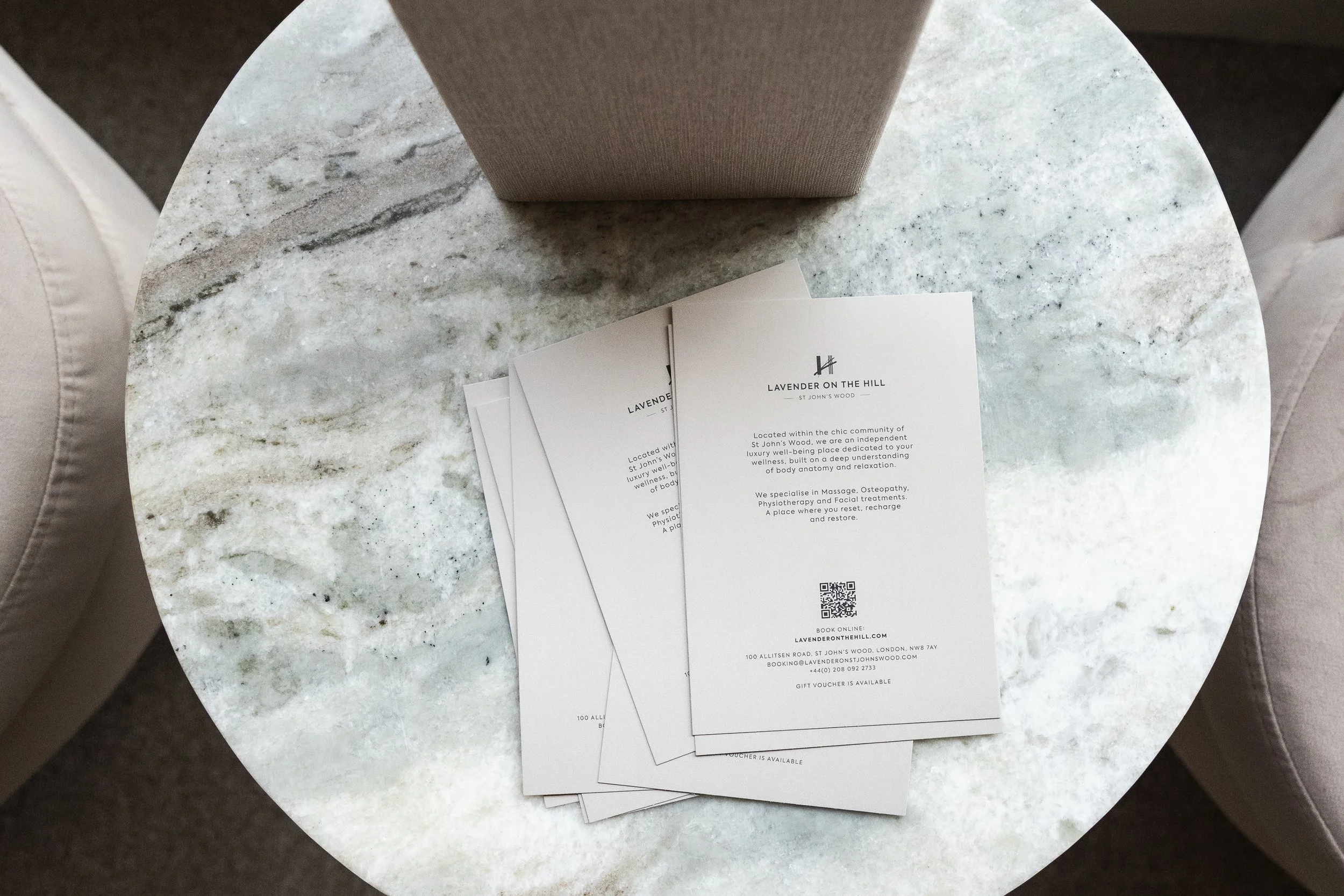 A round, gray marble table with several white pamphlets or flyers stacked on it. The flyers are for Lavender on the Hill, located in St. John's Wood, London.