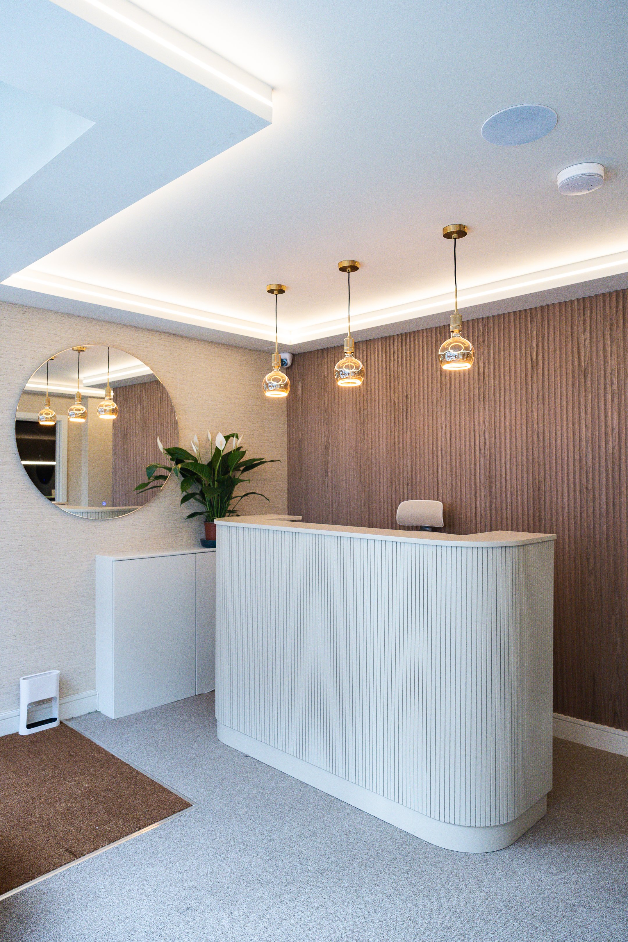Modern hotel or office reception area with a curved white front desk, three pendant lights, a plant, a round mirror on the wall, and neutral-colored walls and carpet.
