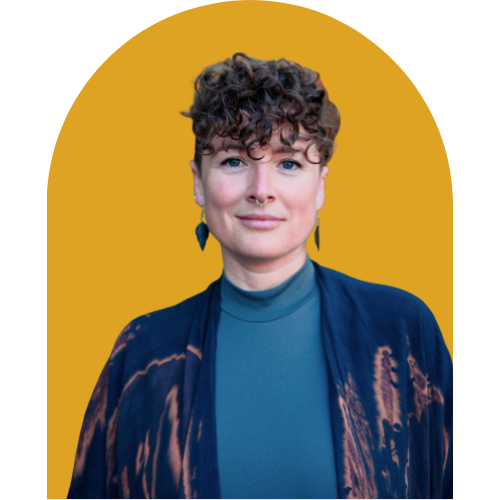 A woman with short curly hair, light skin, and a septum piercing, wearing a blue top and patterned jacket, poses against a yellow background with a partial black shape.