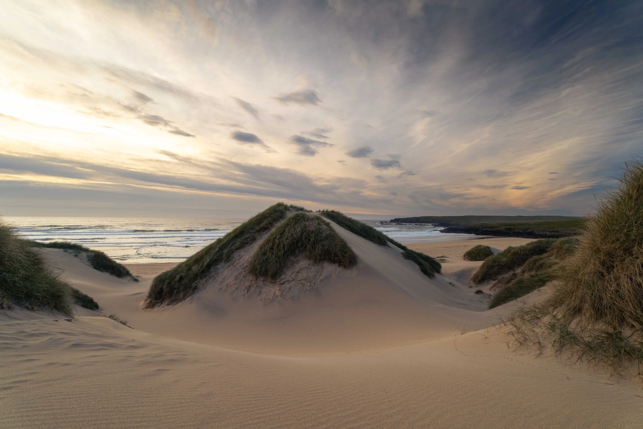 Sandy beach with sand dunes covered in grass, ocean waves, and a cloudy sky during sunset.