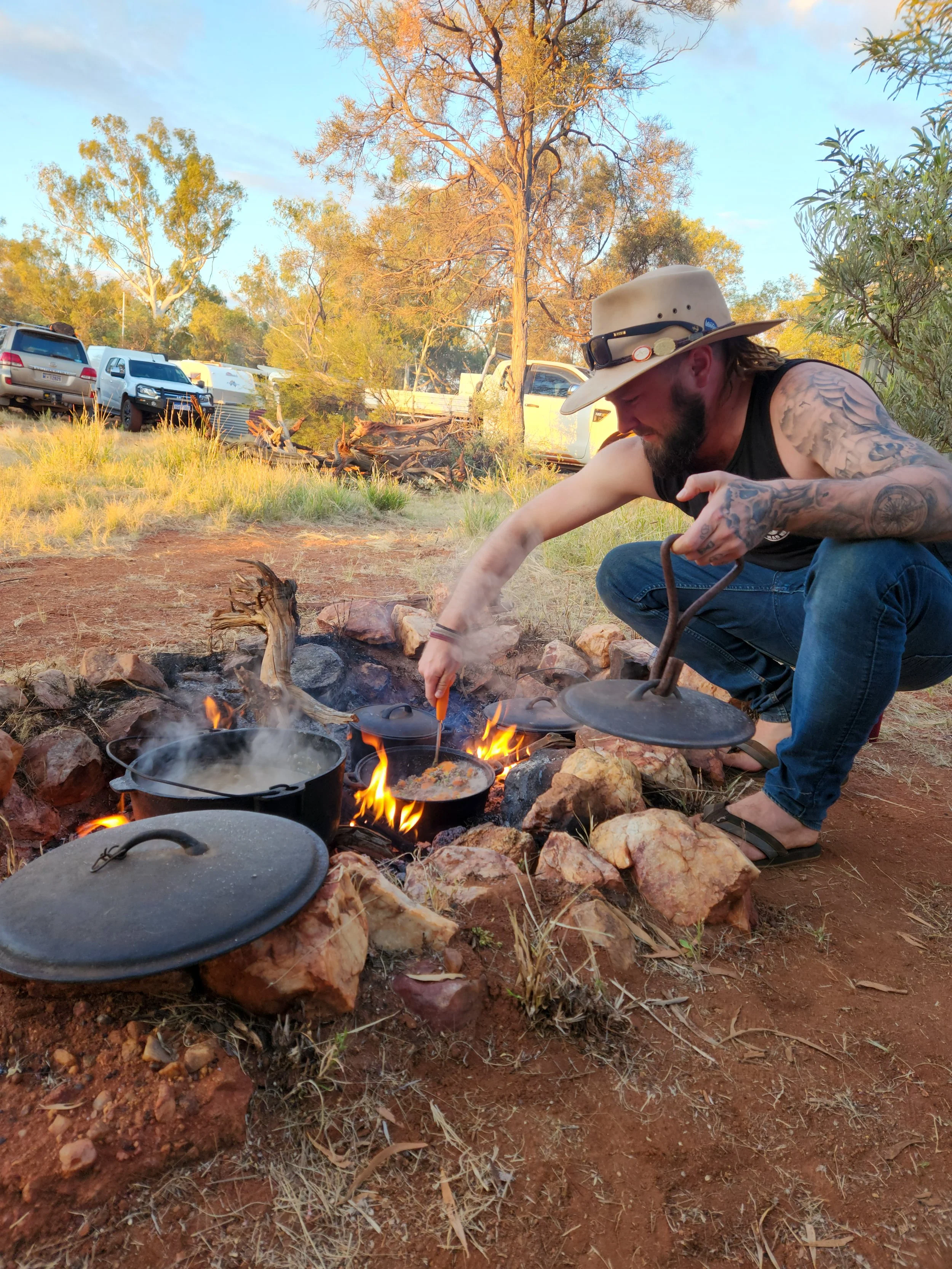 A man with tattoos, wearing a wide-brimmed hat and black sleeveless shirt, squats over an open campfire with three cast iron pots cooking. The outdoor setting is a dry, grassy area with trees and parked cars in the background.