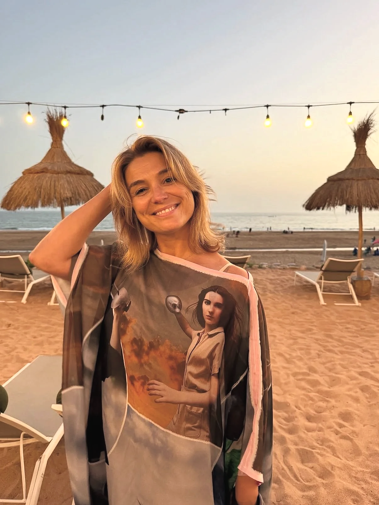 Een lachende vrouw op het strand bij zonsondergang, gekleed in een kleurrijke jas met een afbeelding van een vrouw met een balsak in de hand.