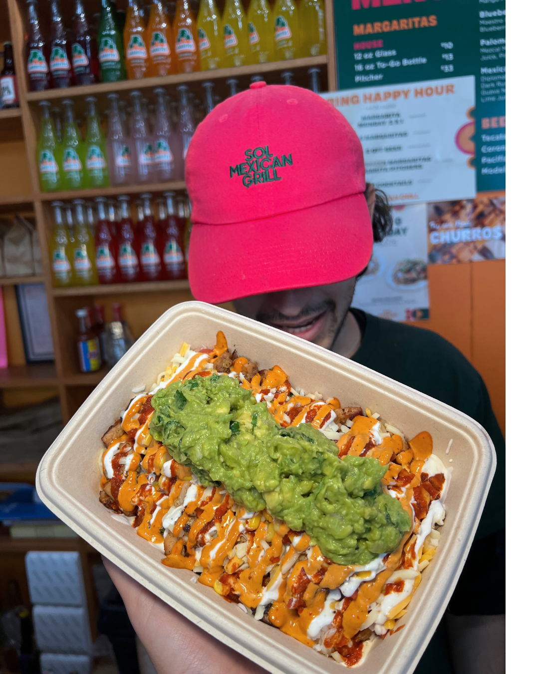 A person wearing a red cap with 'Sol Mexican Grill' embroidered on it holds a takeout container filled with Mexican food, topped with guacamole and drizzled with sauces. The background shows shelves with bottles of soda and a menu board.