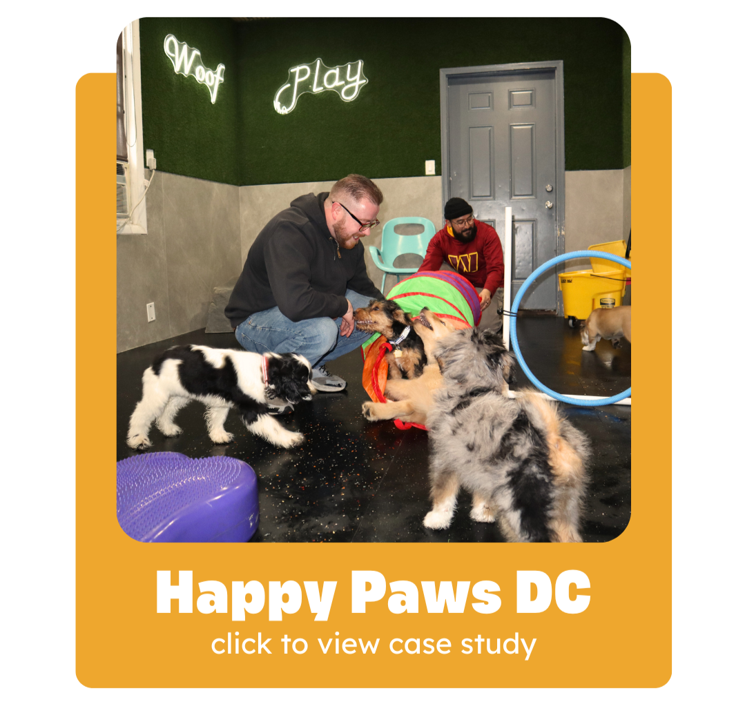 Two men playing with several dogs inside a dog training or playroom, with neon signs that say 'Woof' and 'Play' on the green wall background.