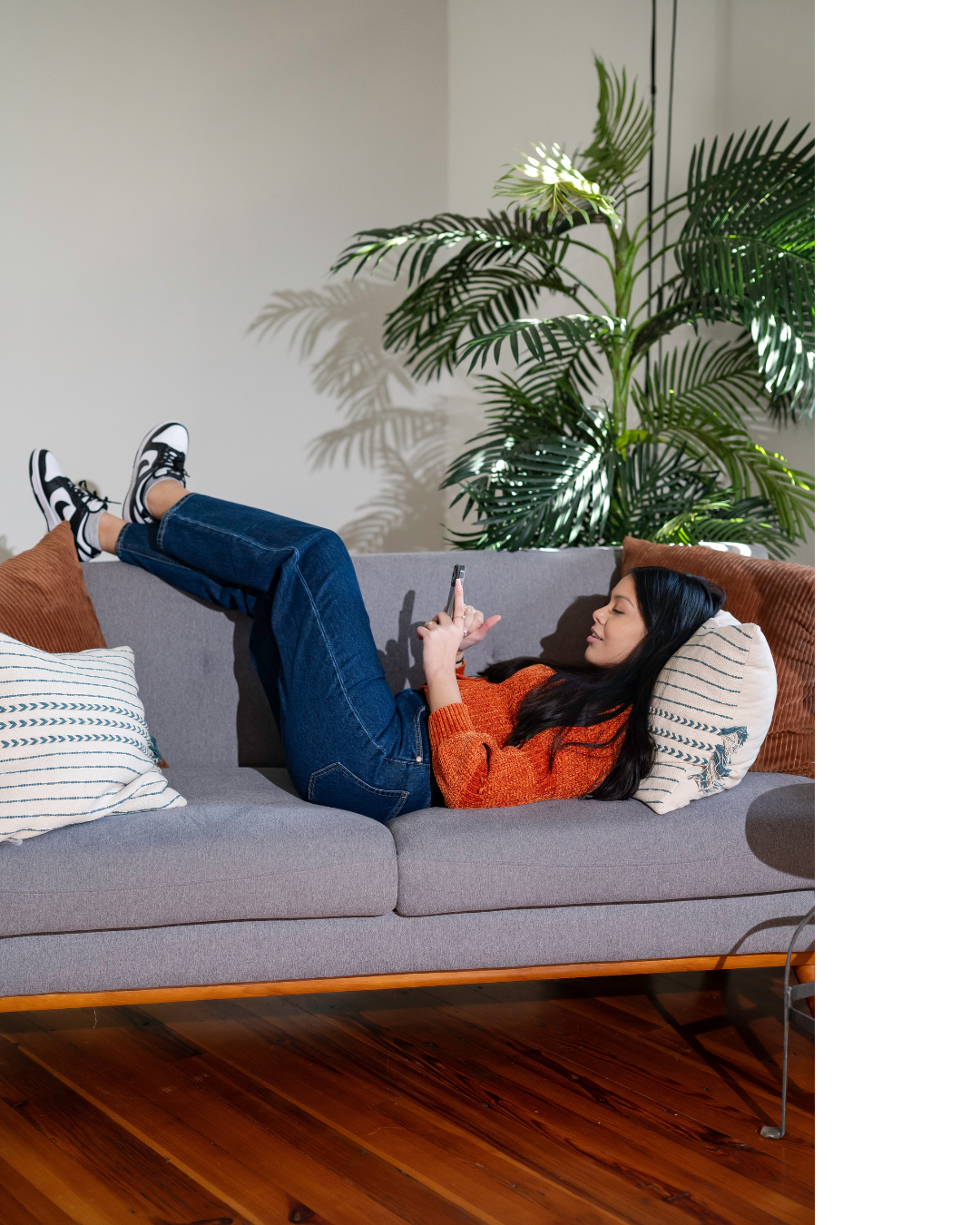 A woman lying on a gray sofa, using her phone, surrounded by throw pillows, with a large green indoor plant behind her.