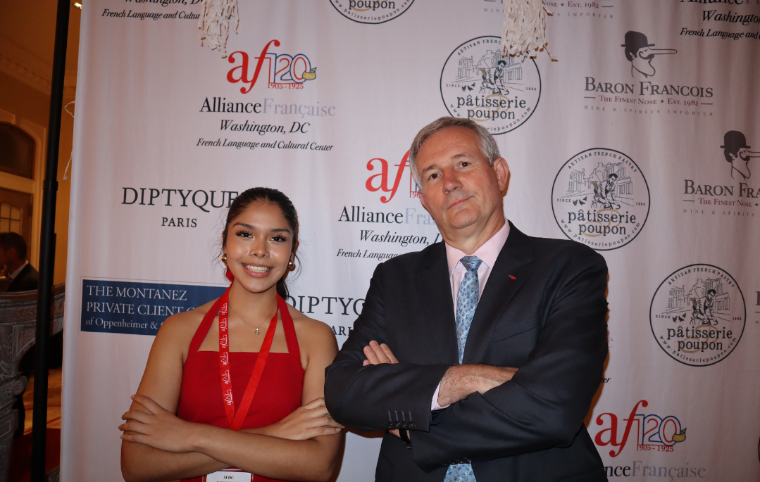 A young woman and an older man stand in front of a backdrop with logos and text, arms crossed, at a formal event. The woman wears a red dress and a red lanyard, smiling, while the man wears a dark suit with a light pink shirt and blue tie, with a serious expression.
