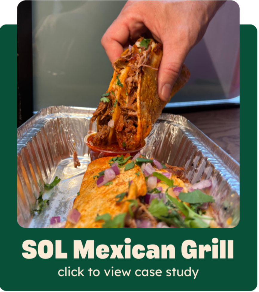 A hand holding a shredded beef taco with vegetables, topped with cilantro and onions, served with another taco with meat and toppings in a foil tray.