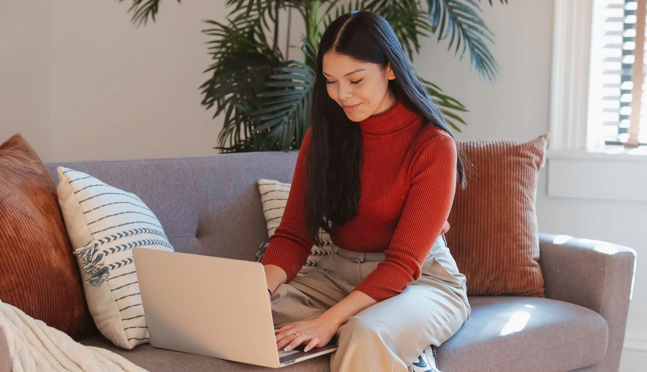 A woman with long dark hair wearing a red turtleneck sweater and beige pants sitting on a gray sofa using a silver laptop, with striped and textured cushions behind her, in a cozy living room with a large plant and a window with blinds.
