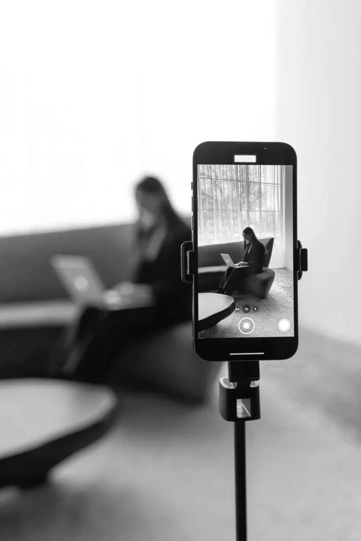 A smartphone mounted on a tripod capturing a woman sitting on a chair with a laptop, near a large window.