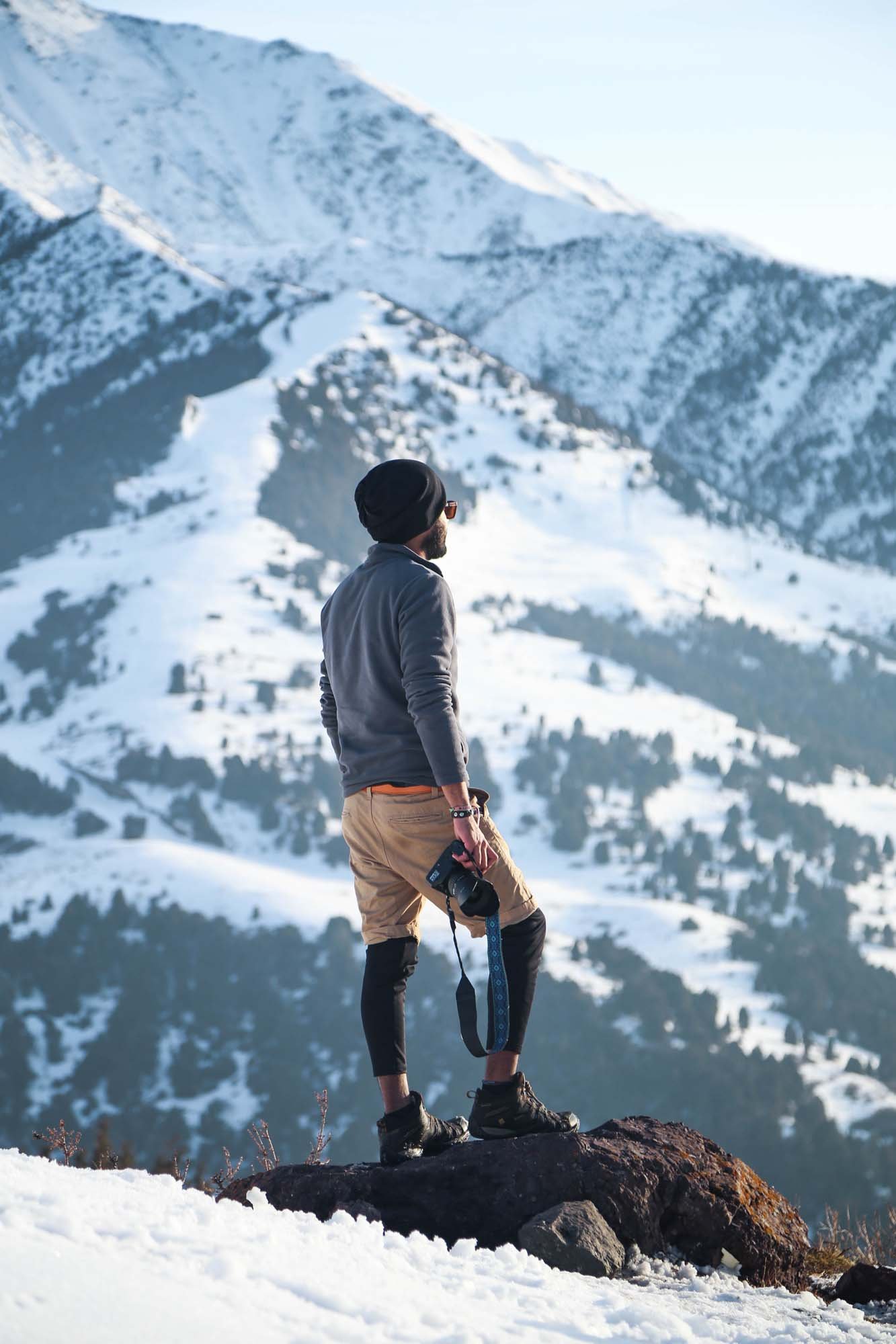 A man standing on a rock in a snowy mountain landscape, holding a camera, wearing outdoor gear, with snow-covered peaks in the background.
