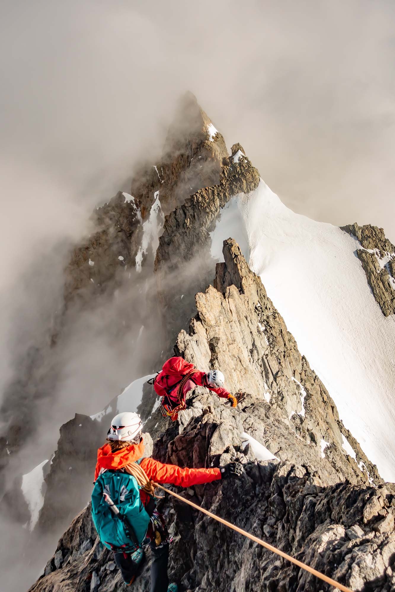 Two mountain climbers wearing helmets and safety gear ascend a rugged, snow-capped mountain ridge with clouds surrounding the peak.