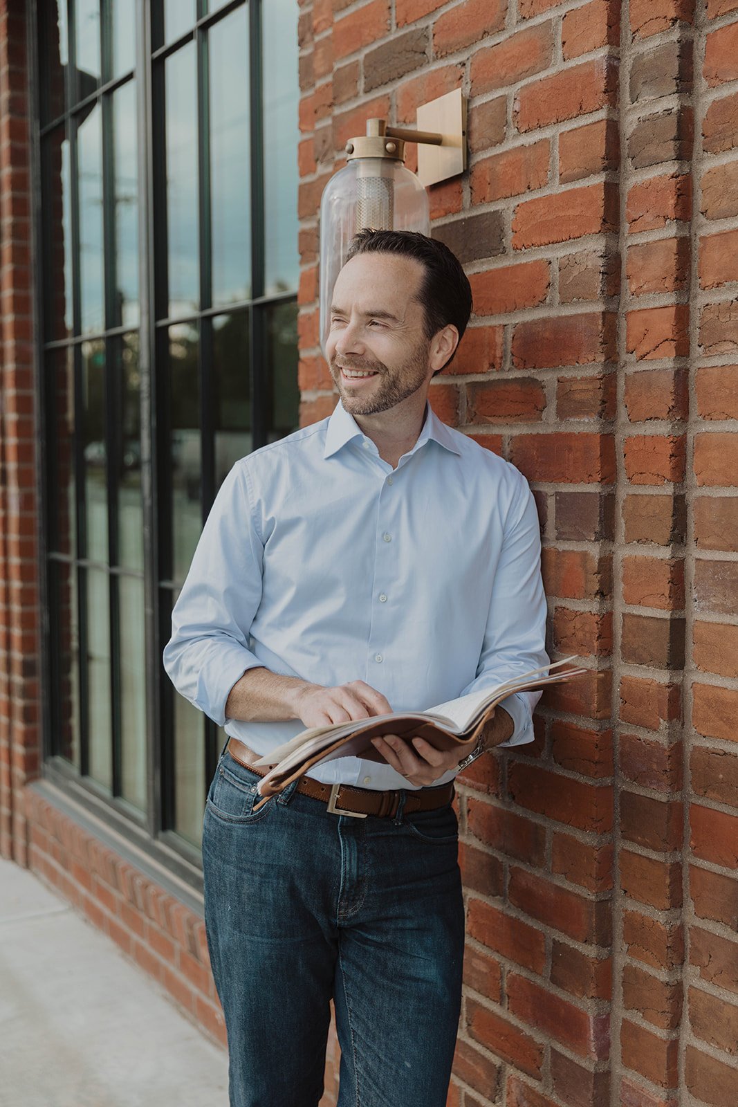 A man with dark hair and a beard, wearing a light blue dress shirt, stands outside leaning against a red brick wall, holding an open book and smiling.